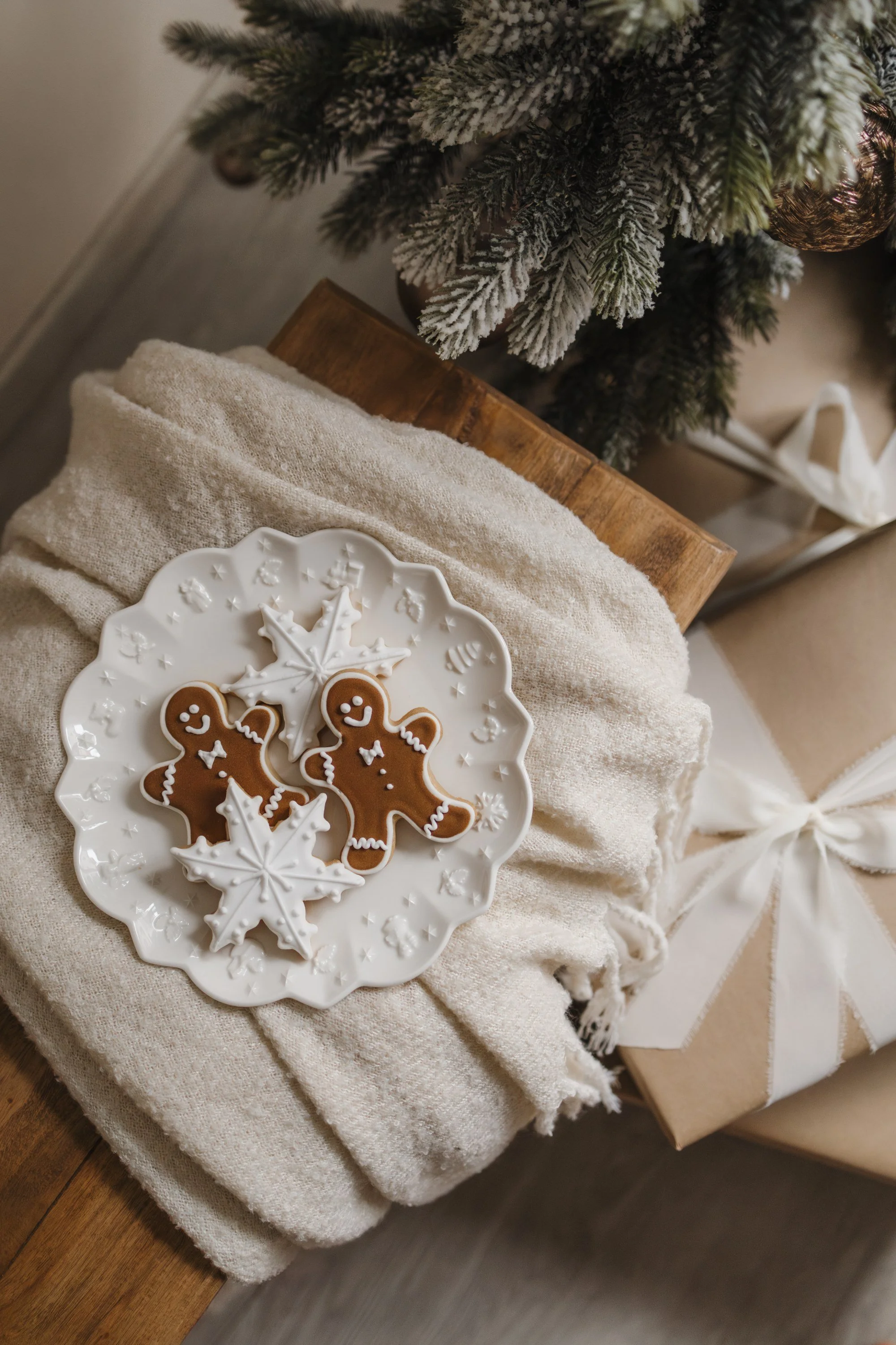 Christmas cookies on a white decorative plate with snowflake and gingerbread man shapes, placed on a cozy beige blanket near a Christmas tree with snow-dusted branches.