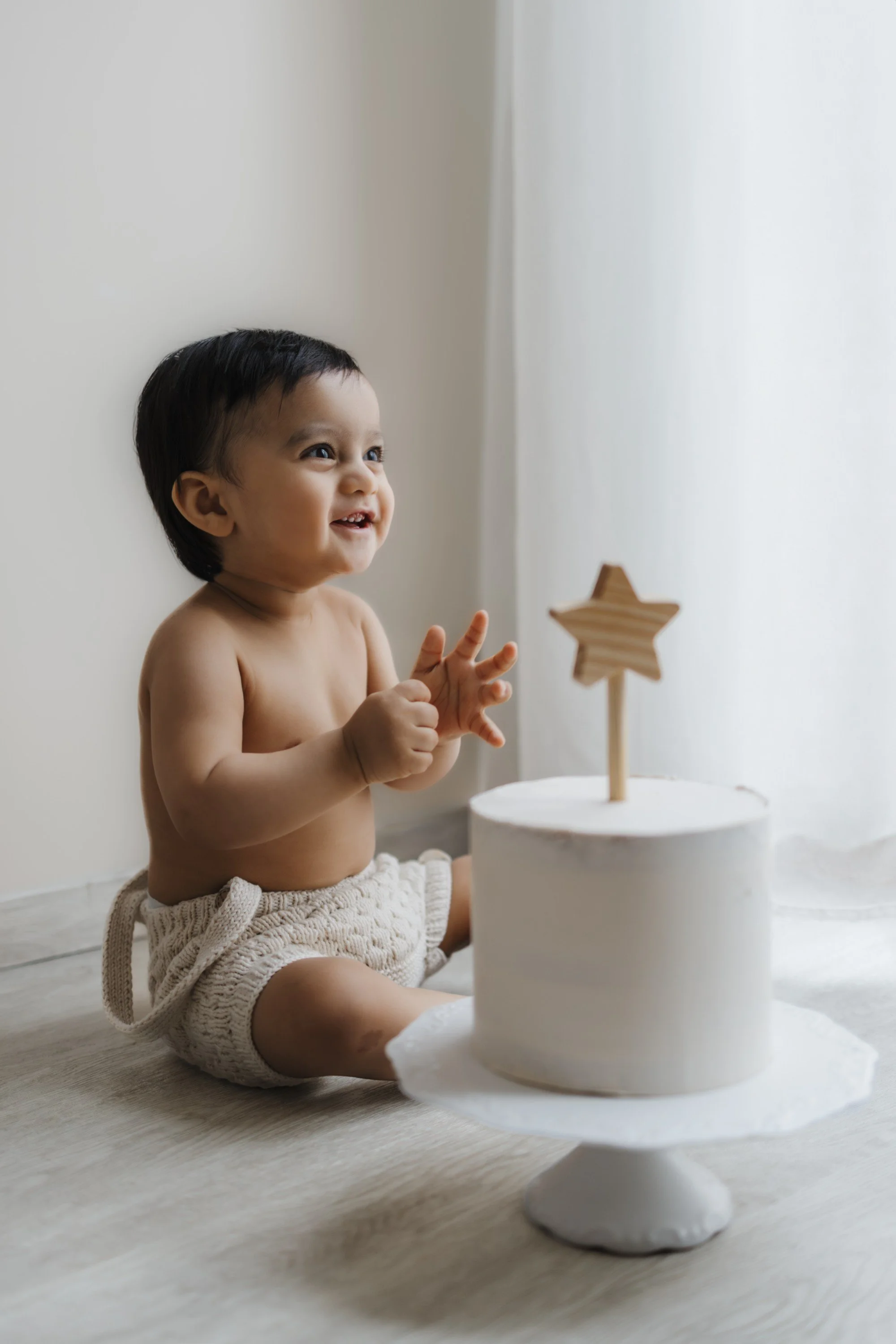 A woman with long, wavy hair is gently kissing a toddler on the head while holding him, sitting on a small wooden stool in a bright room with a white curtain.