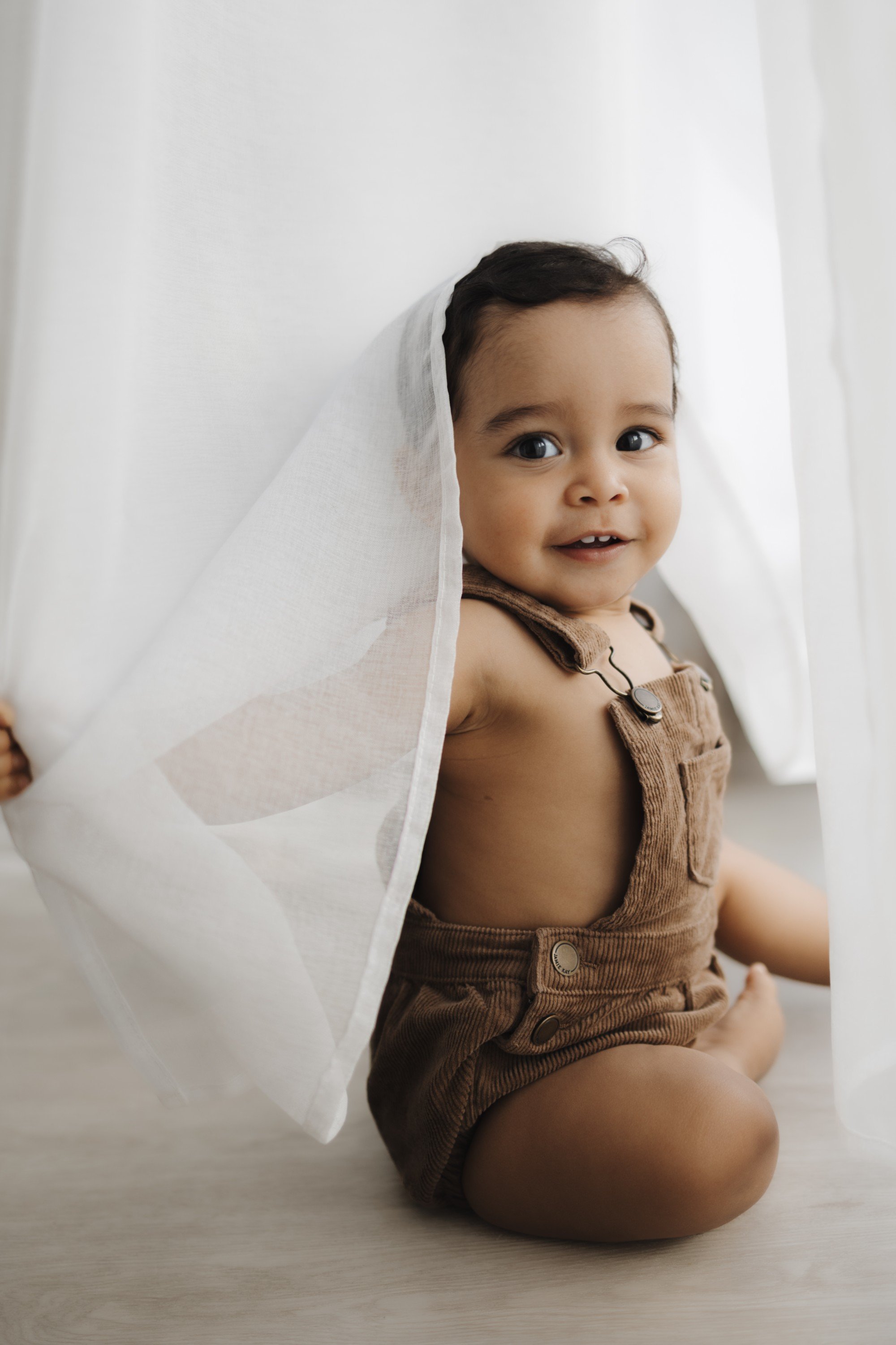 A happy baby sitting on the floor, holding a sheer white curtain, wearing brown overalls, with dark hair and big eyes.