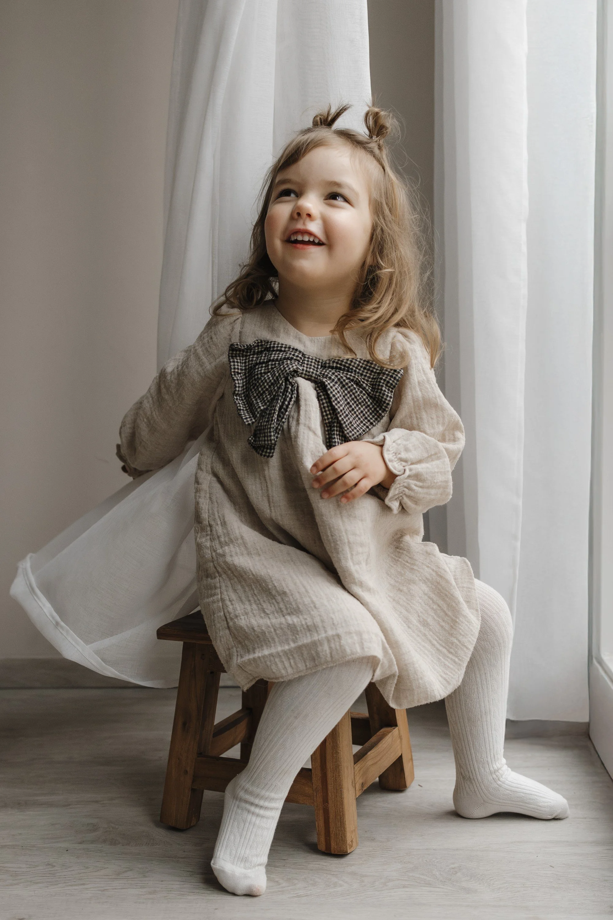 A smiling young girl sitting on a small wooden chair near a window with white curtains, wearing a beige dress with a black and white checkered bow, white tights, and enjoying a bright day.