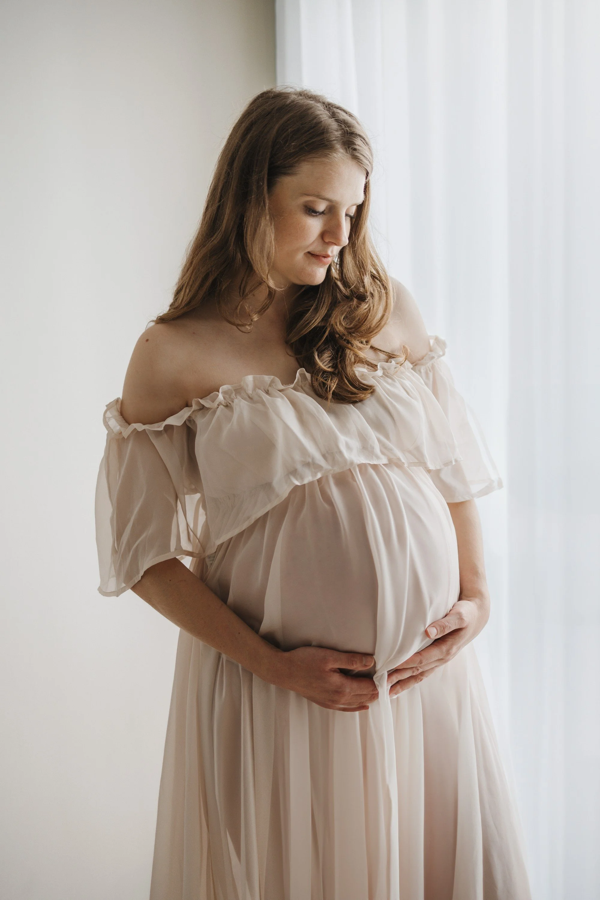 Pregnant woman in a soft-colored dress holding her belly and looking down gently.