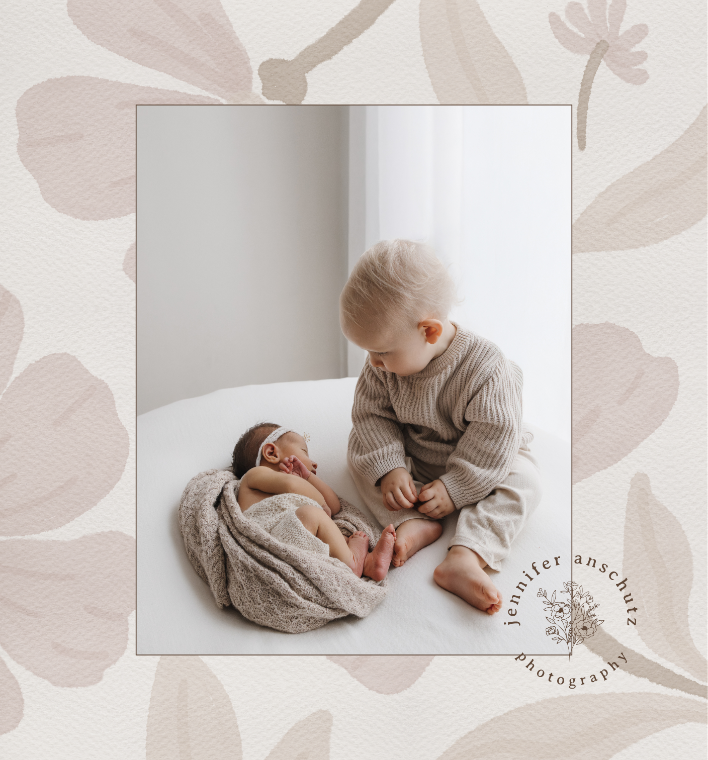 Two young children, a newborn and an older sibling, sitting on a white bed in a softly lit room. The baby is wrapped in a beige blanket with a headband, and the older child is leaning over, looking at the baby, dressed in a beige sweater and cream pants. The background features light-colored curtains and a minimal, neutral room design.