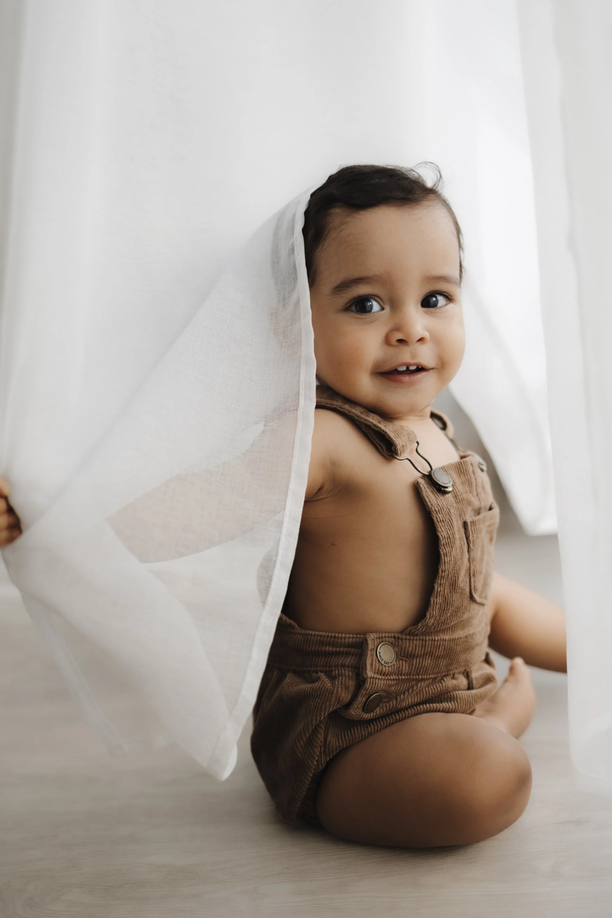 A smiling young toddler with dark hair and big eyes, sitting on a light-colored floor, partially covered by a sheer white curtain.