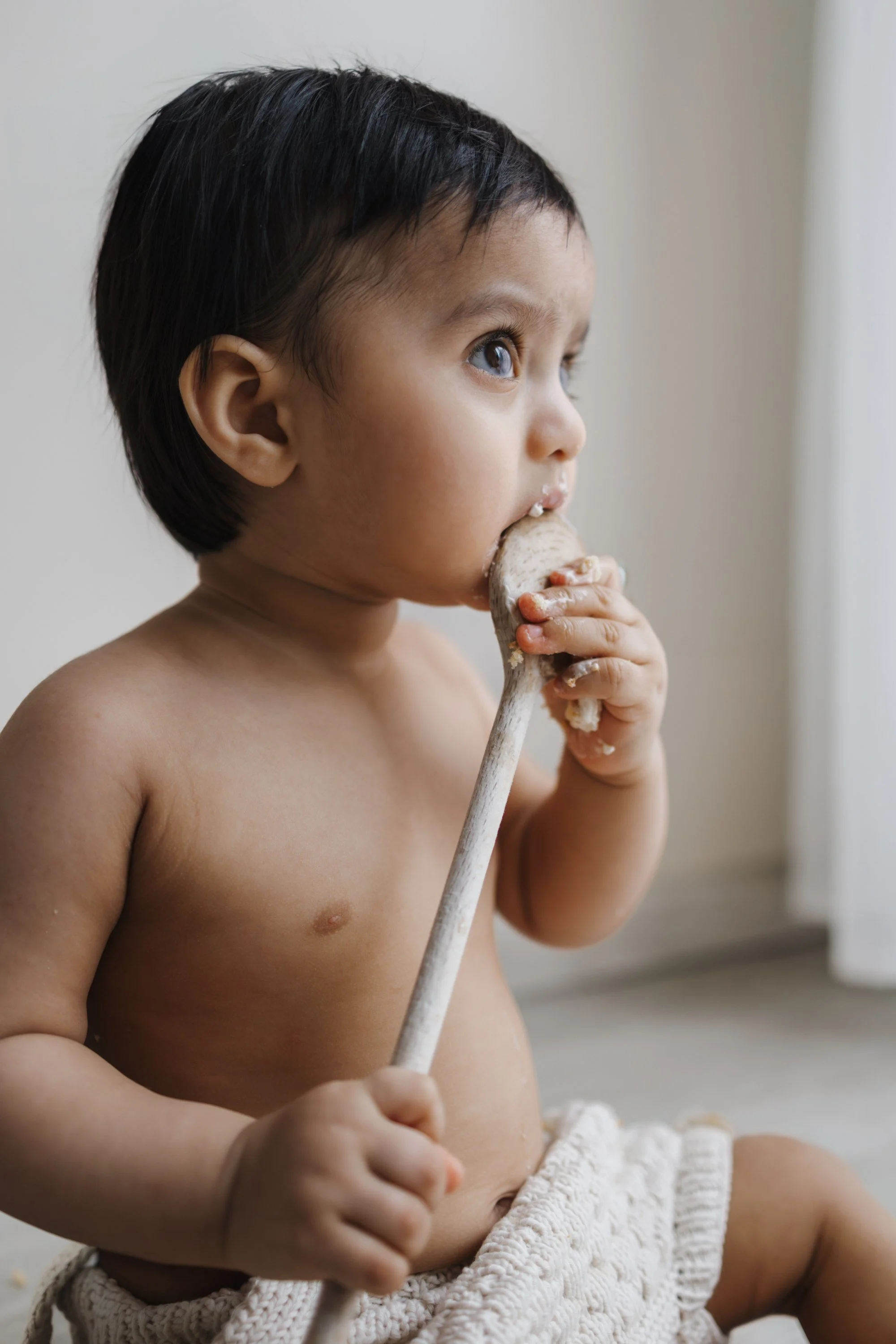A young child with dark hair and brown skin is sitting shirtless, holding a wooden spoon and eating something with their hands.