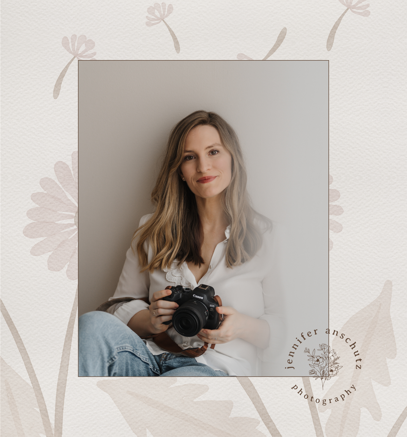 A woman with long, wavy hair holding a camera, sitting against a light-colored wall, smiling gently.
