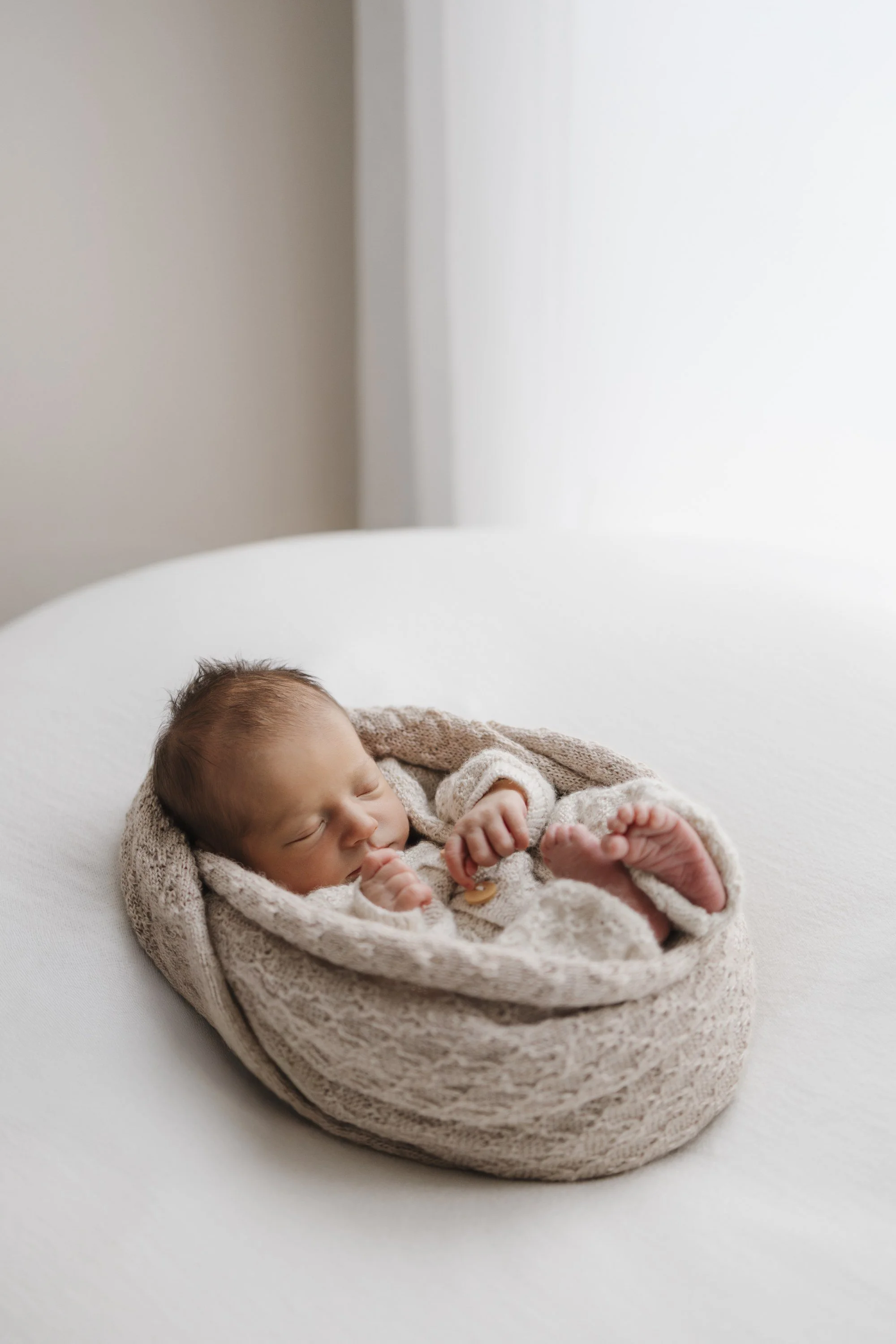 A sleeping newborn baby wrapped in a knitted beige blanket on a white surface.