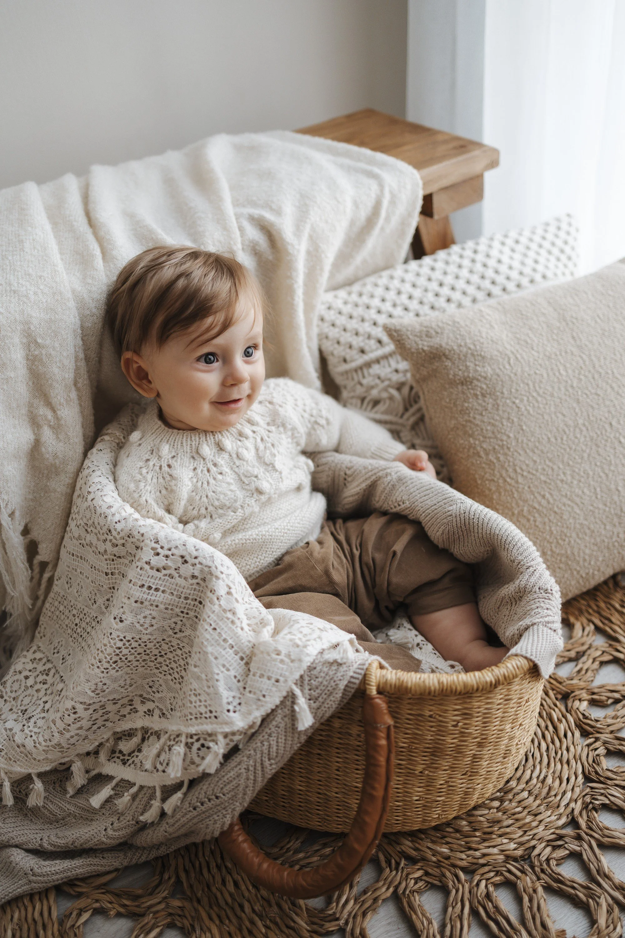 A young child sitting in a wicker basket on a woven rug in a cozy, neutral-toned living room with cushions and a throw blanket.