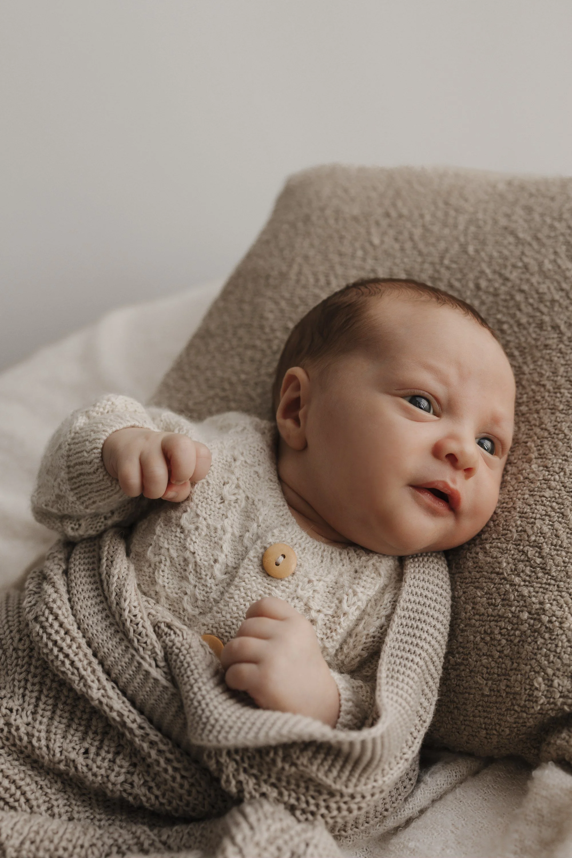 A baby lying on a bed, resting on a plush pillow, wearing a beige knitted sweater with wooden buttons, looking at the camera with a slight smile.