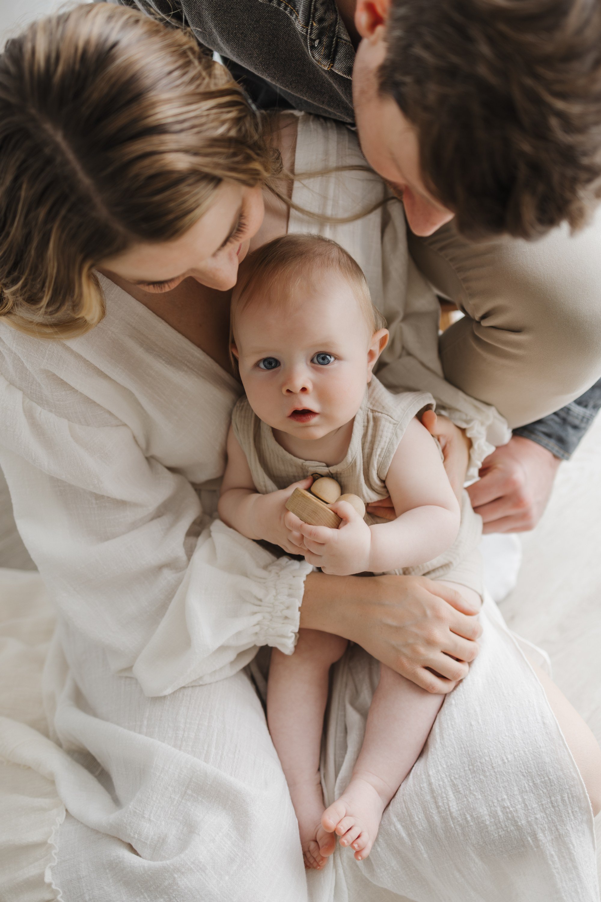 A baby with blue eyes sitting on a woman's lap, holding a wooden toy, surrounded by two adults leaning over and looking at the baby.