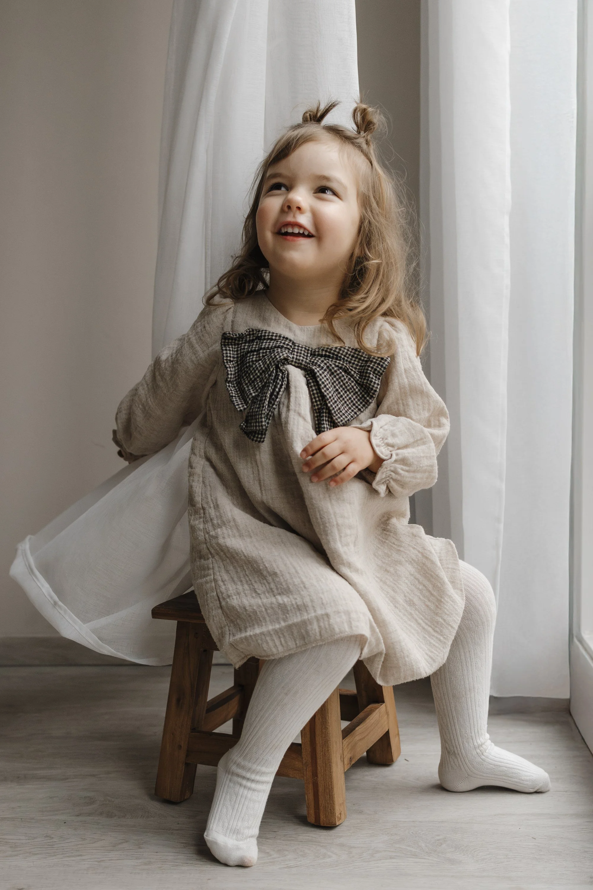A young girl with light brown curly hair styled in two buns, sitting on a small wooden stool near a window with white curtains, wearing a beige dress with a large checkered bow, white tights, and smiling while looking upward.