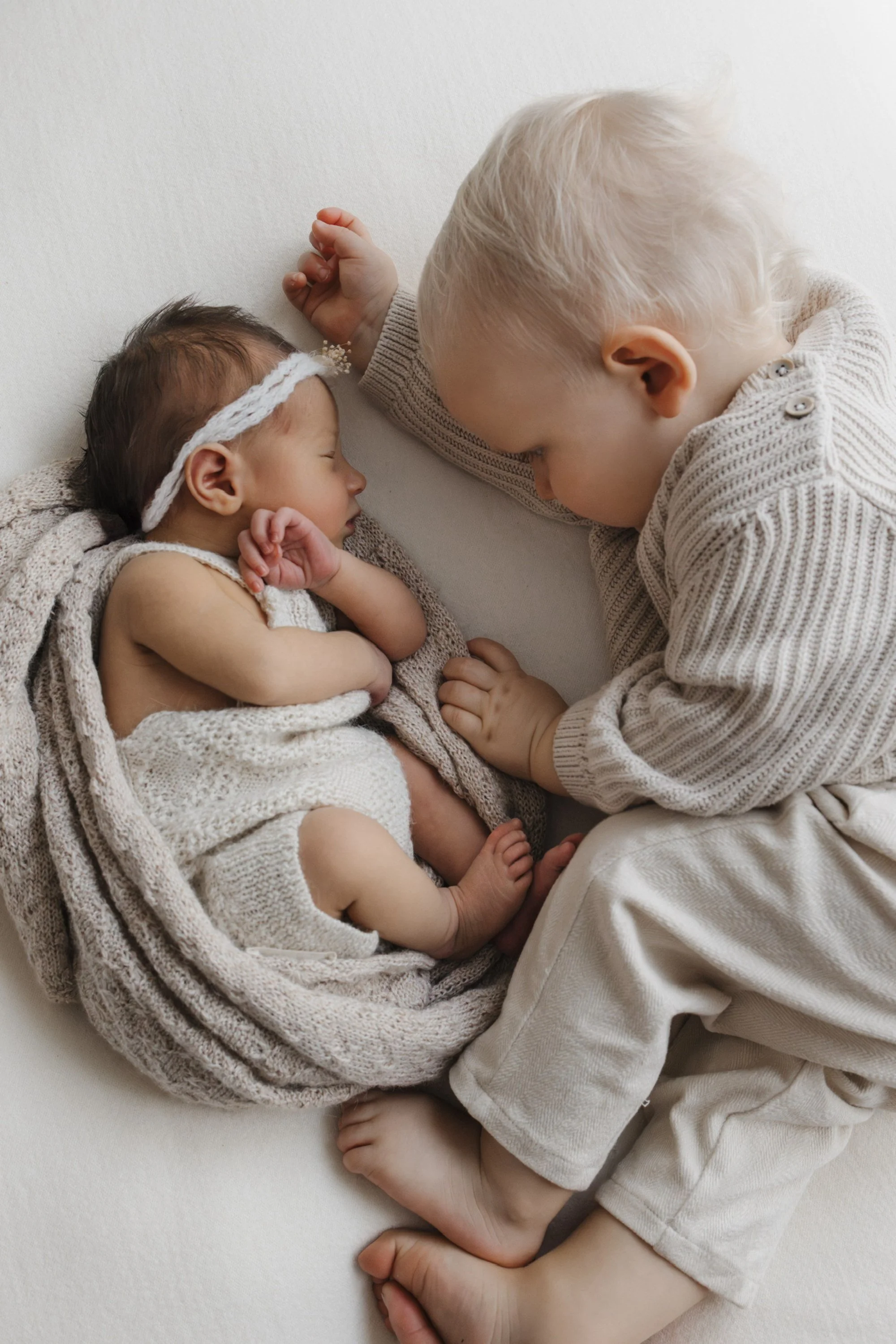 A young blonde-haired boy and a newborn girl are lying on a white surface, facing each other. The girl is wrapped in beige knit clothing with a headband, and the boy is wearing a beige sweater and pants. They are touching foreheads in a tender, intimate moment.