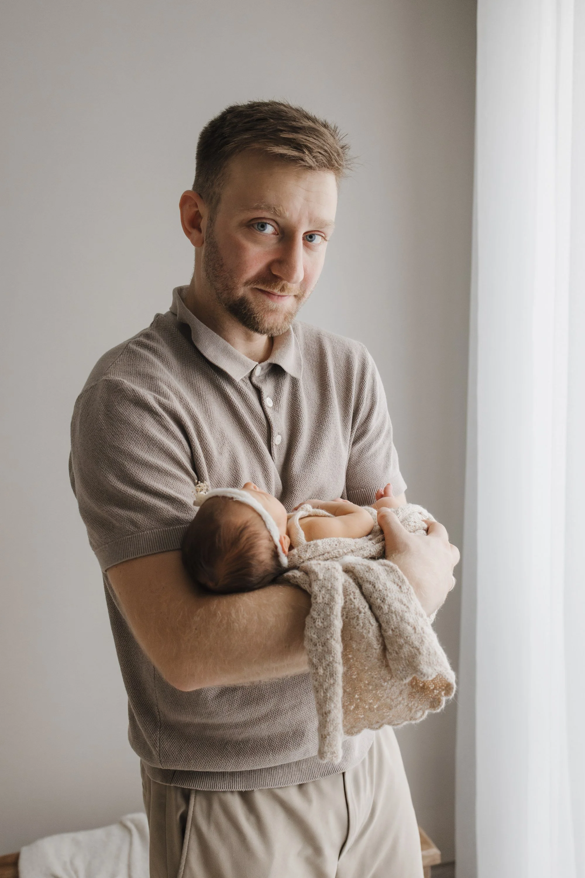 A man is holding a baby wrapped in a blanket near a window.