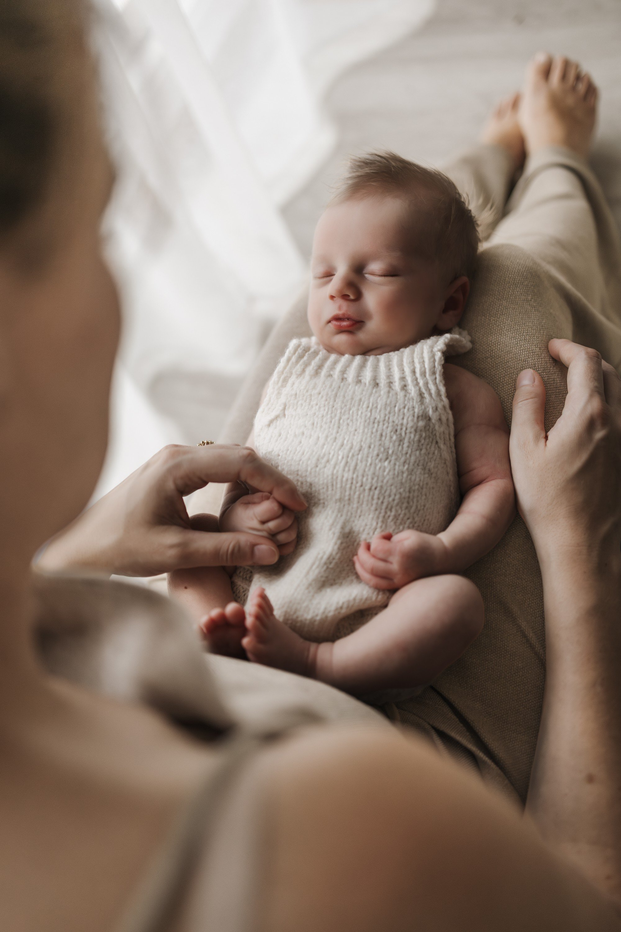 A newborn baby sleeping peacefully on a person's lap, with hands gently holding the baby's hand and arm.