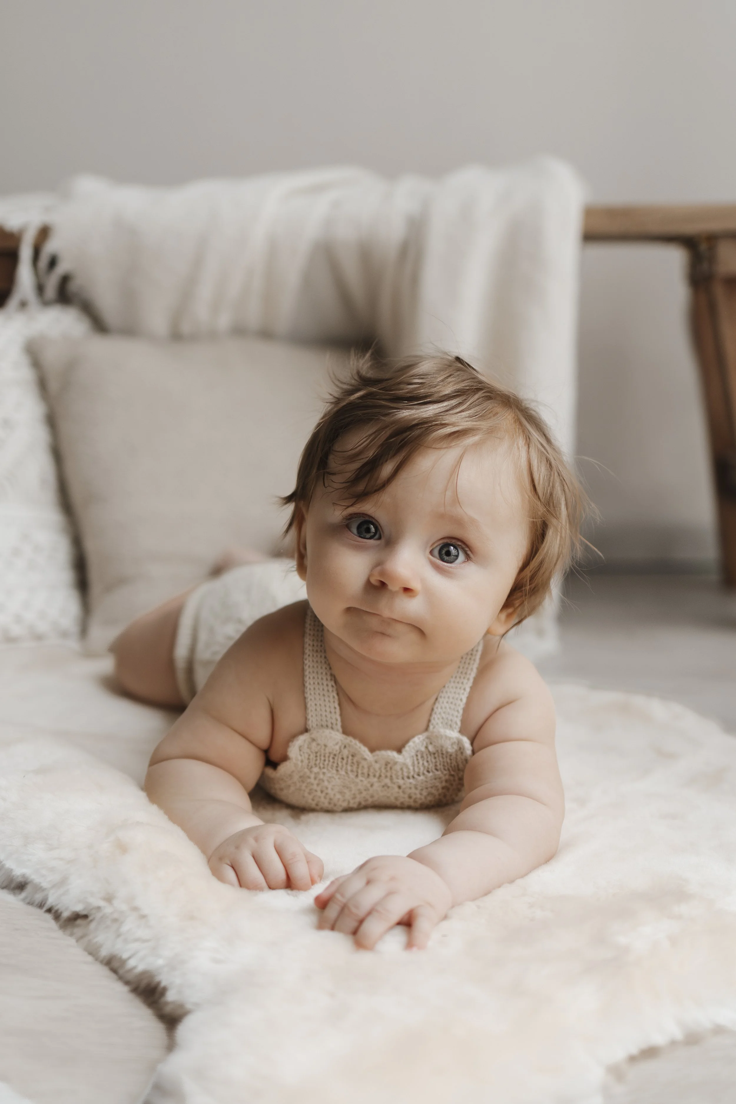 A baby lying on its stomach on a soft, cream-colored blanket with a neutral-colored pillow and a cozy setting in the background.