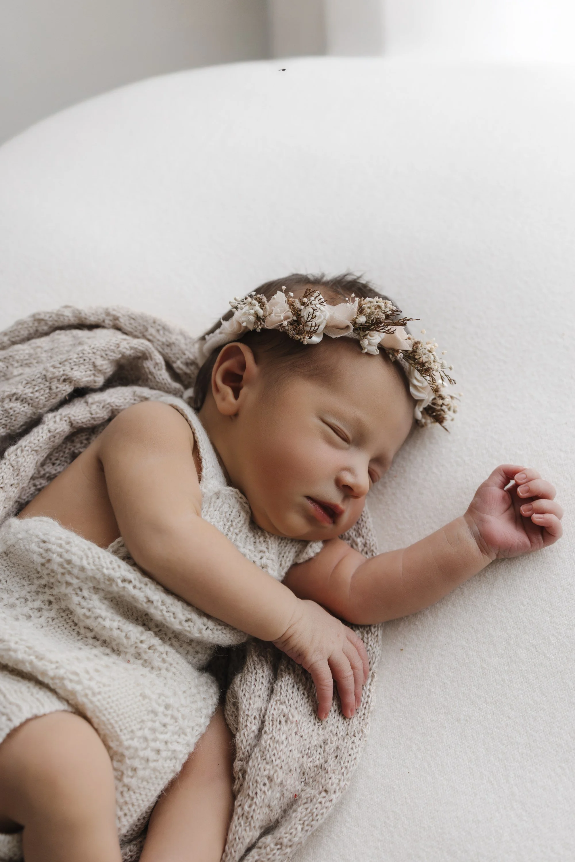 A sleeping baby girl with a floral headband, wrapped in knitted blankets, lying on a soft white surface.