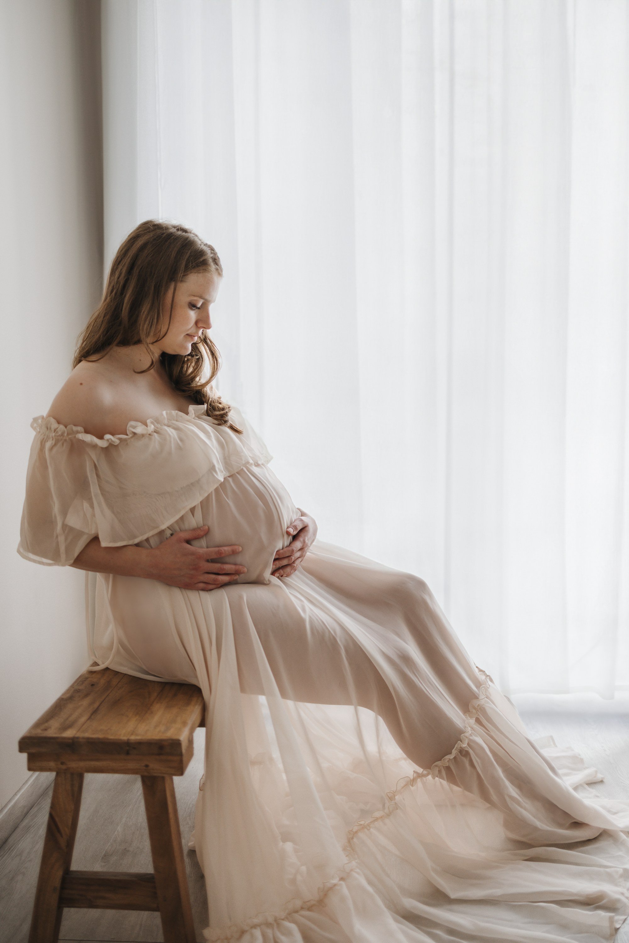 A woman with long, wavy hair holding and kissing a baby boy sitting on a small wooden chair in a bright room with white curtains.