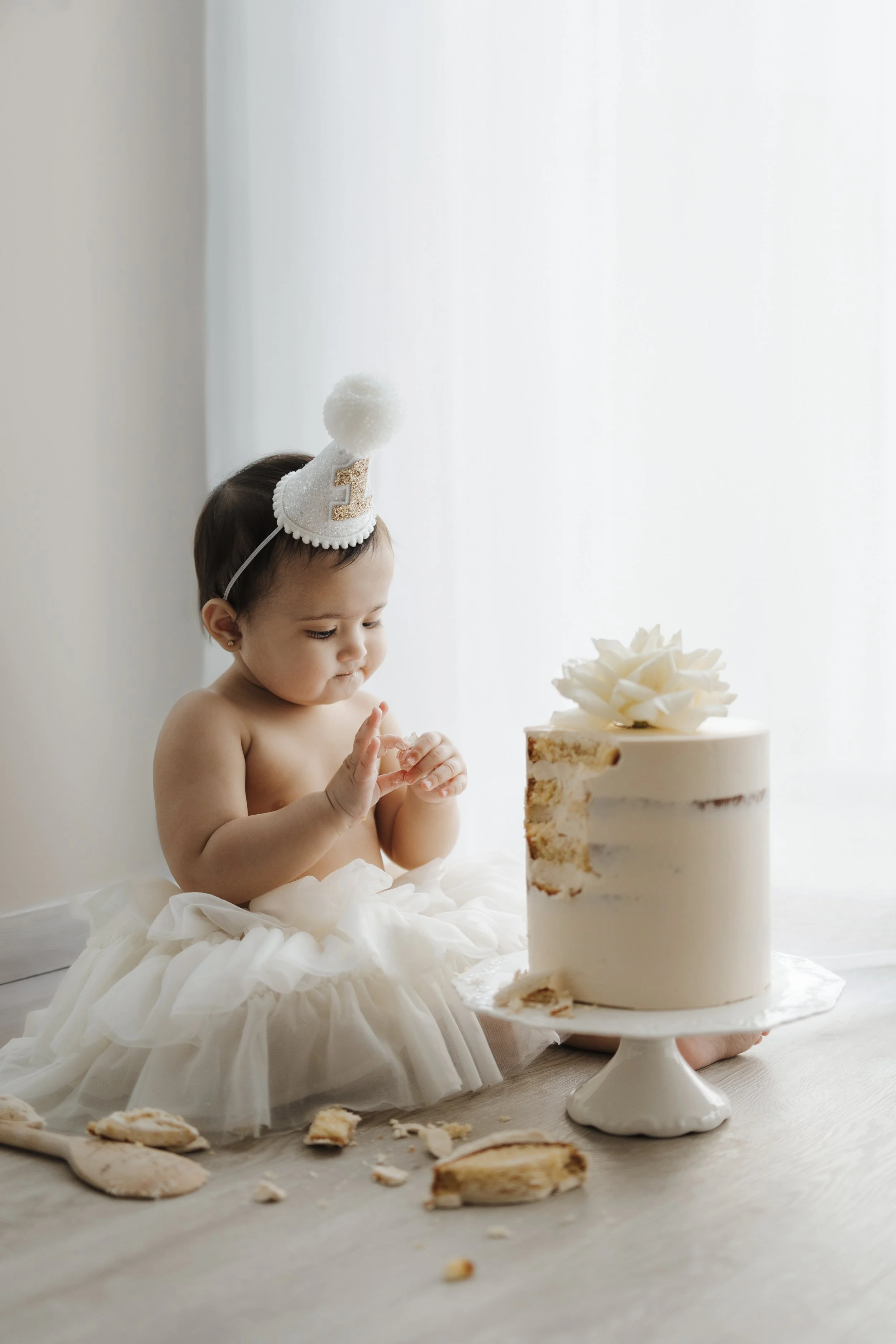 Young toddler girl in a white tutu dress with a birthday hat, sitting on the floor next to a partially eaten cake with white icing, a large flower decoration on top, and cake crumbs scattered around.