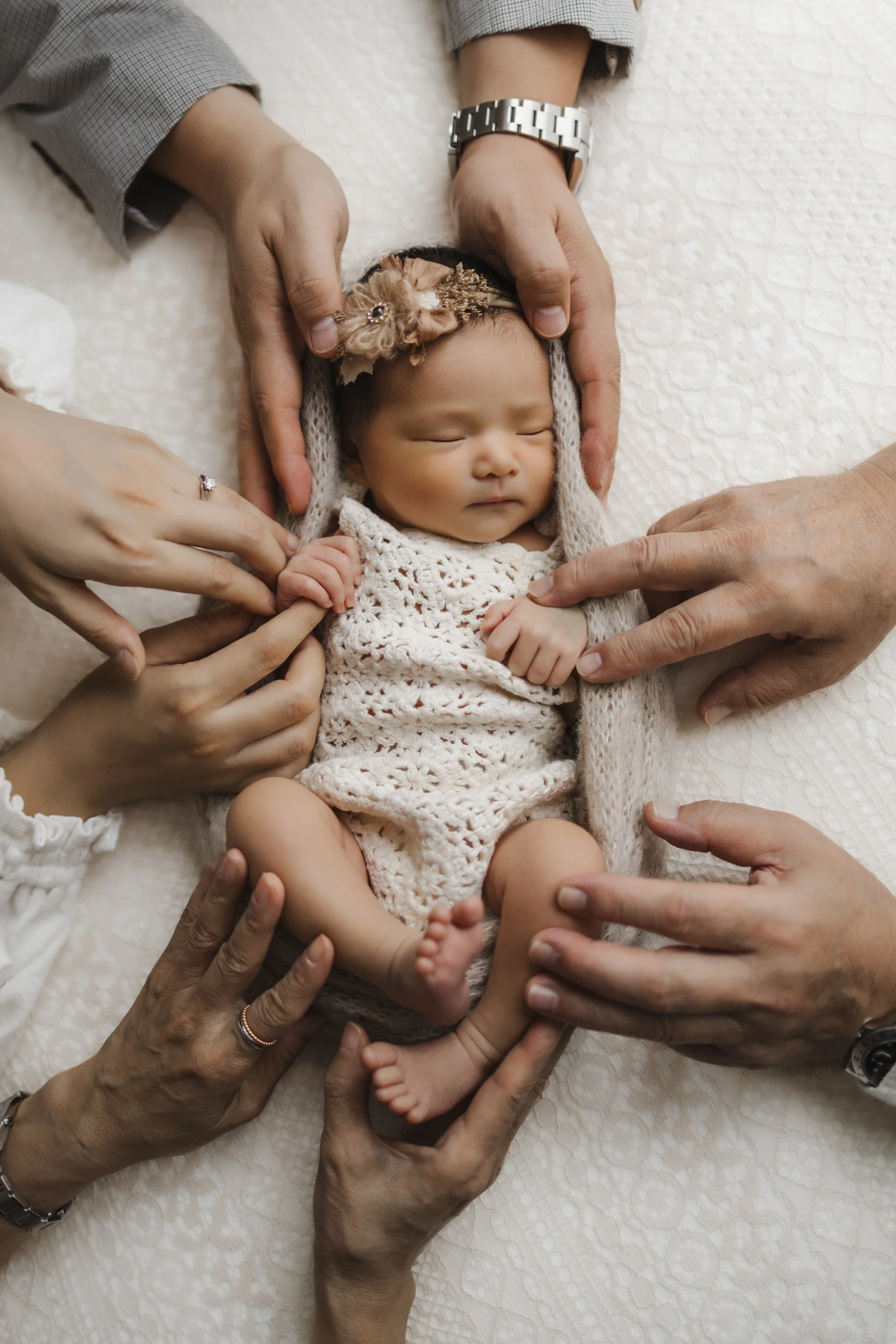 A newborn baby wearing a cream crocheted outfit and a headband with a flower, lying on a soft cream blanket surrounded by multiple adult hands gently supporting and holding the baby.