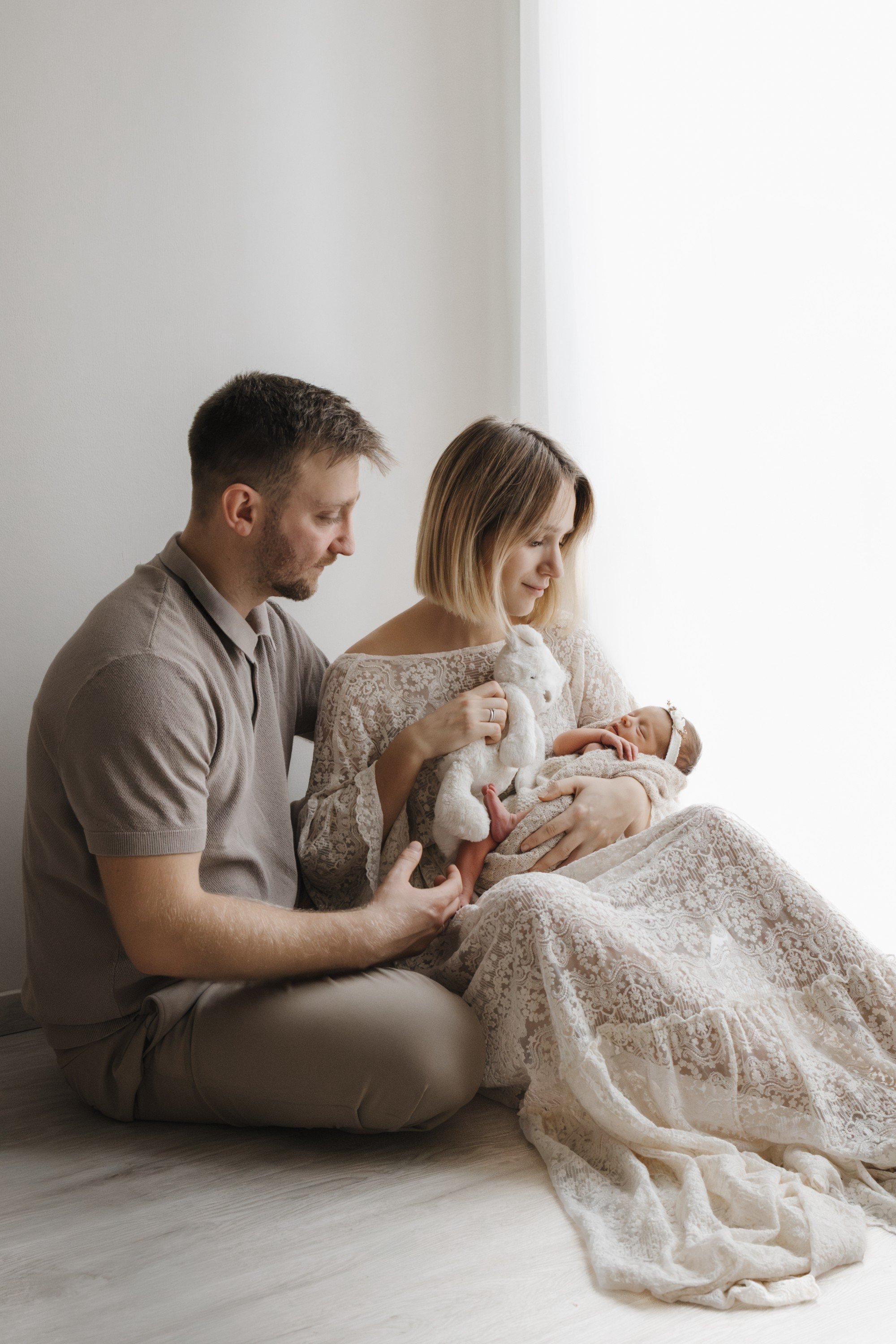 A couple sitting on the floor holding their newborn baby by a window with natural light, the woman dressed in lace and holding a stuffed bunny.