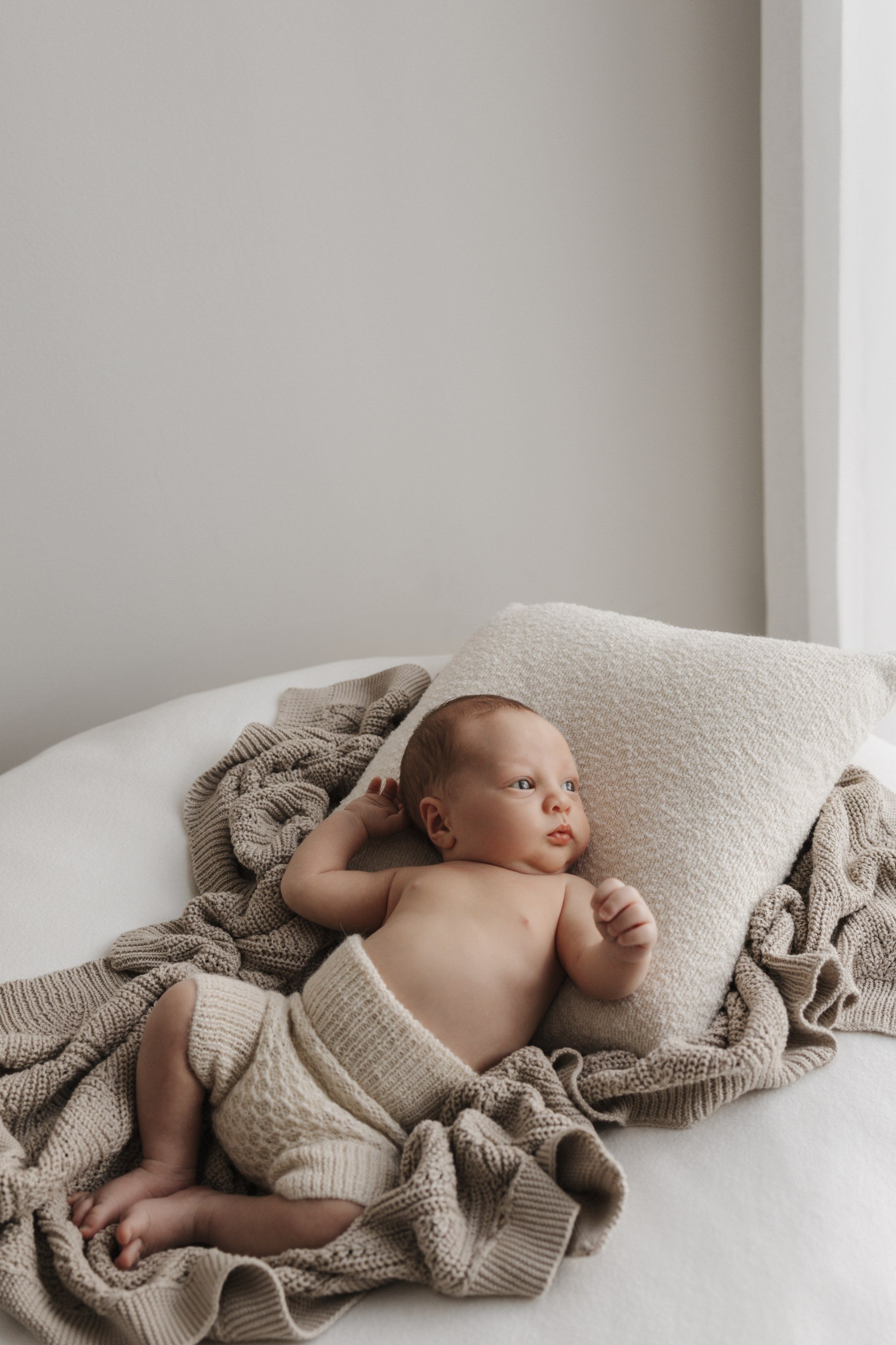 A baby lying on a bed surrounded by beige knitted blankets and pillows, looking off to the side.