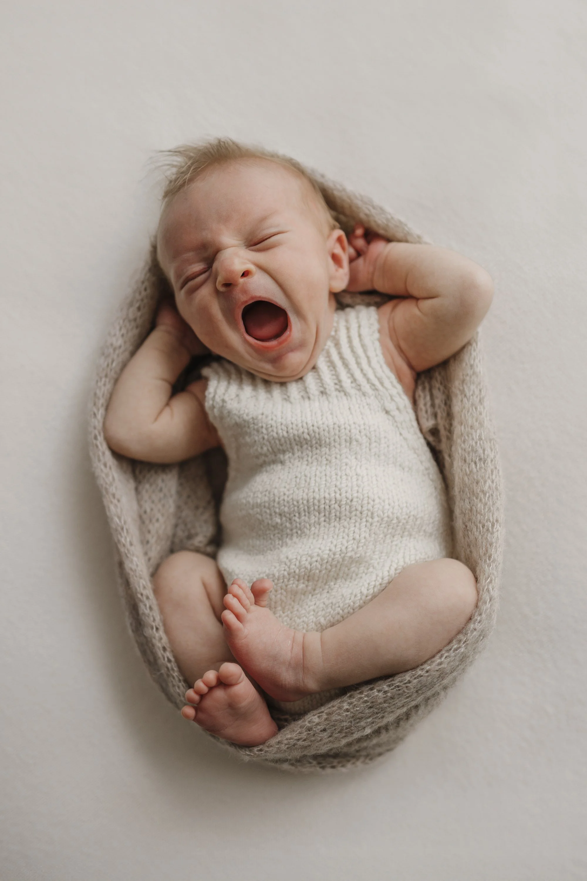 Newborn baby yawning, lying in a beige knitted blanket on a white surface.