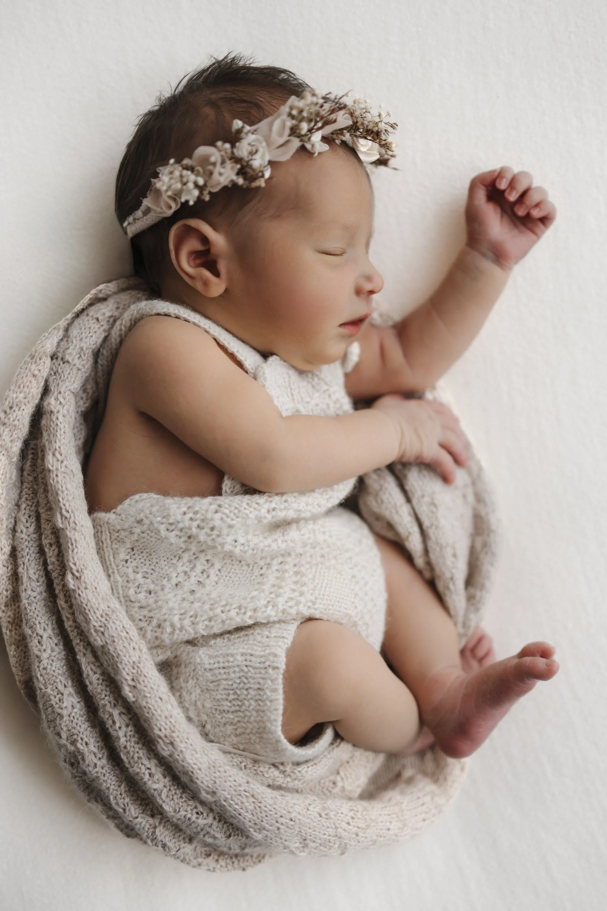 A sleeping newborn baby girl wrapped in a beige knitted blanket, wearing a decorative floral headband, with a peaceful expression and closed eyes, lying on a plain white surface.