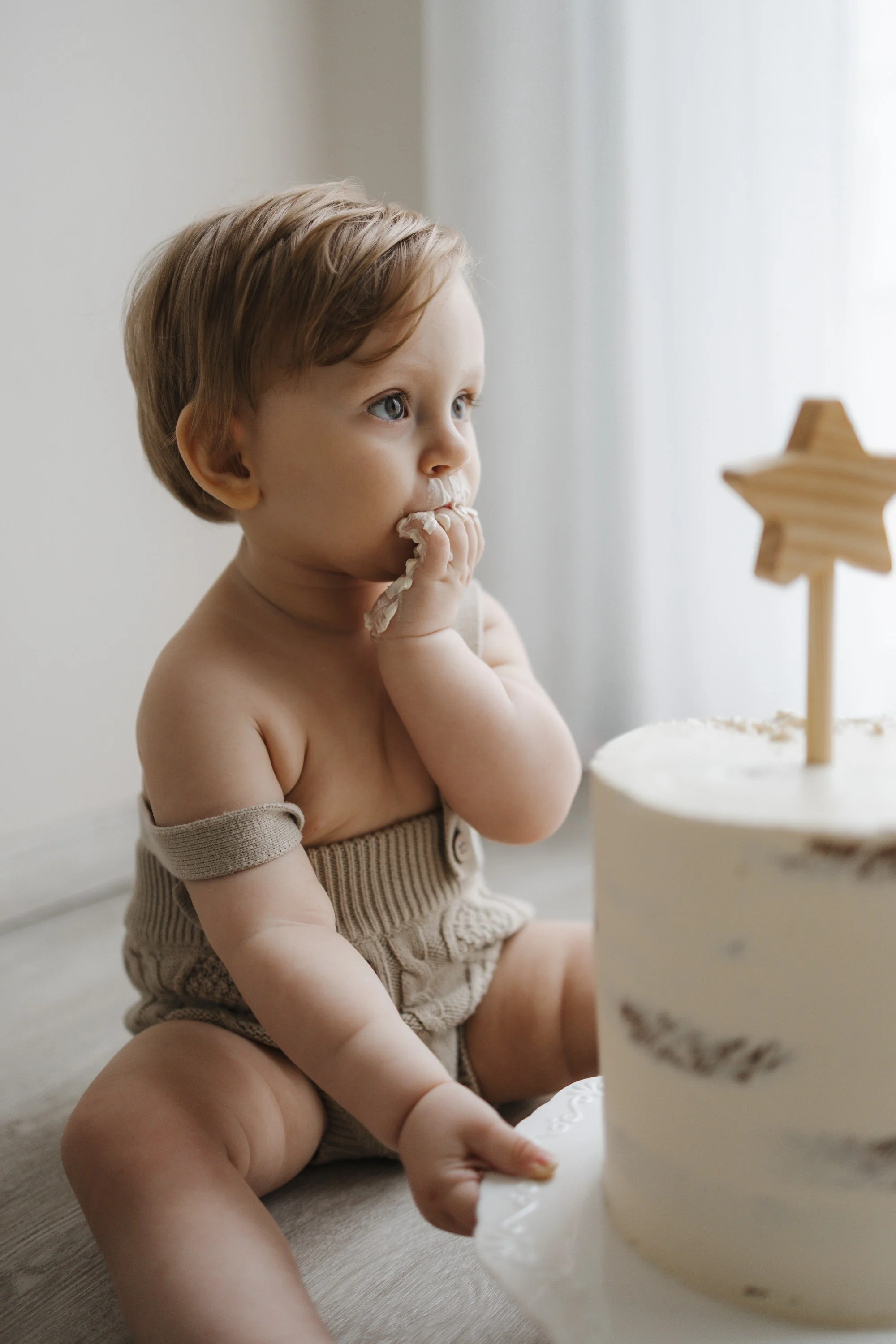 A young child with light brown hair, sitting on the floor, eating cake and looking contemplative next to a white cake with a wooden star topper.