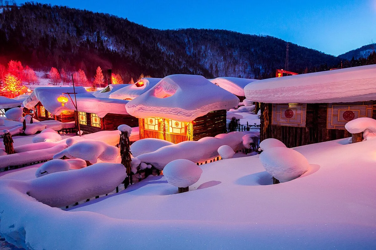 Snow-covered wooden houses with glowing windows and red lanterns in a mountainous village during winter at dusk.
