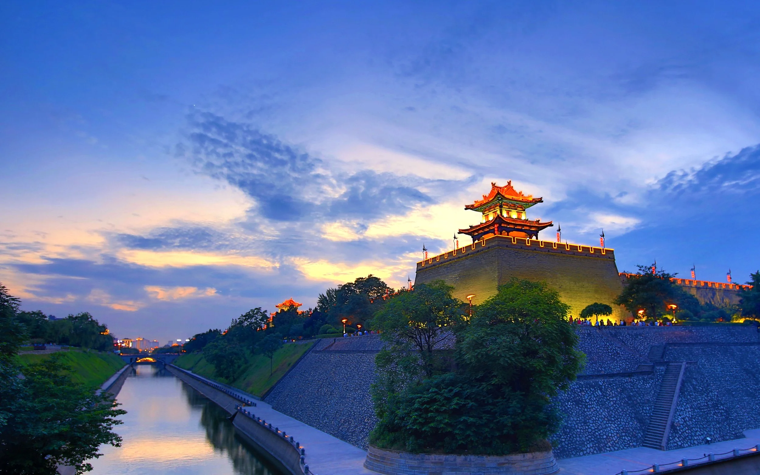 Night view of an illuminated historic Asian castle with a river and city skyline in the background.