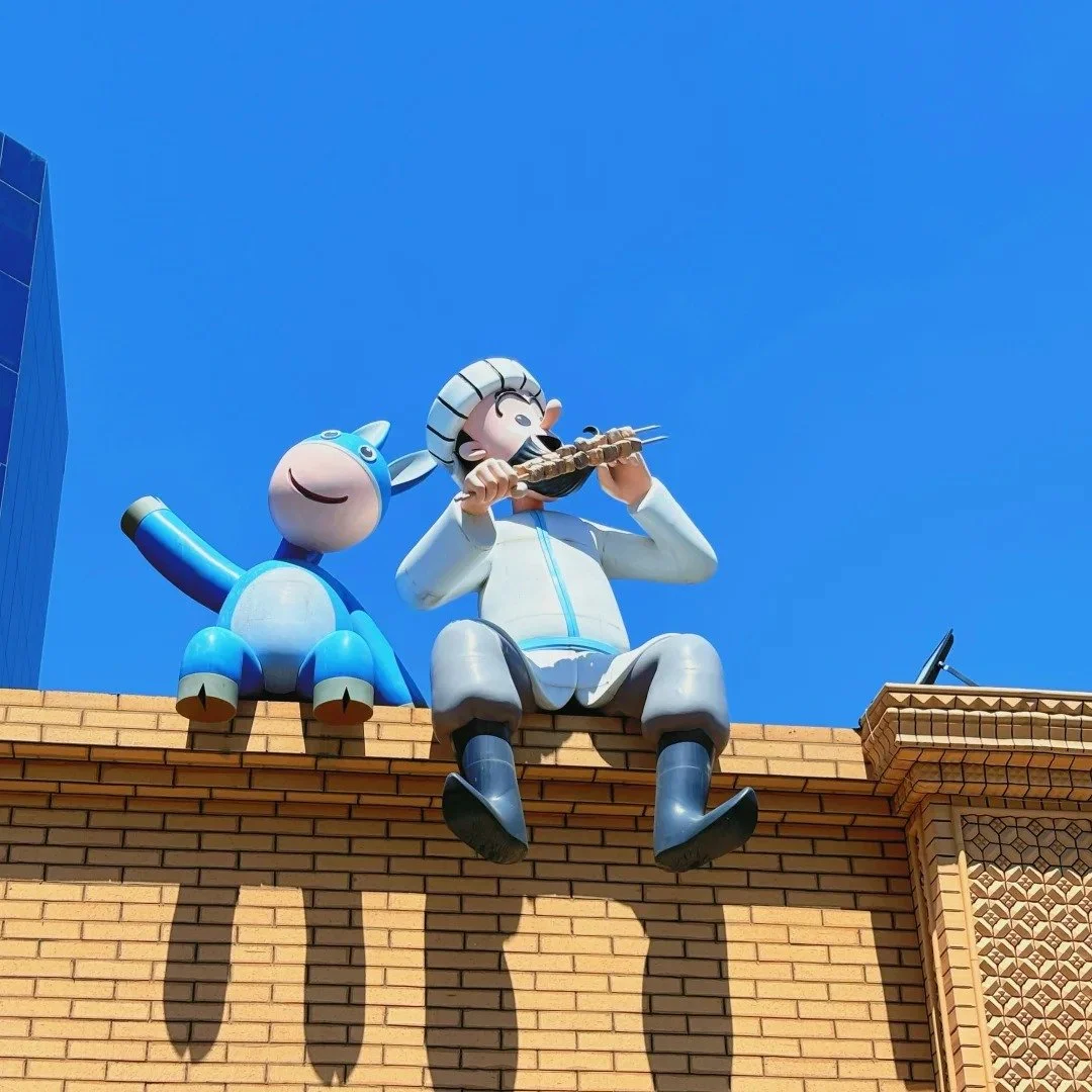 Large cartoon-style statue of a smiling Nasreddin playing a flute, sitting on a building ledge with a blue and white creature beside him, against a clear blue sky. Travel Xinjiang