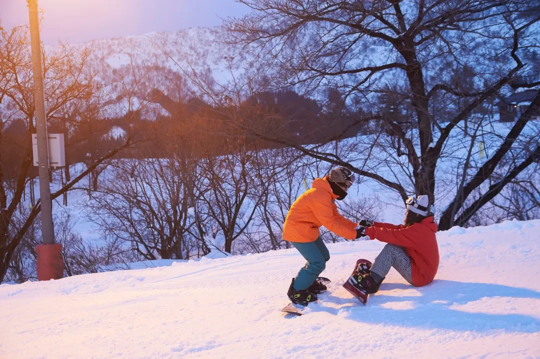 Two children in winter clothes playing in the snow. The scene is set outdoors with snow-covered ground, trees, and a mountainous background. The picture is taken in Snow village, Harbin travel