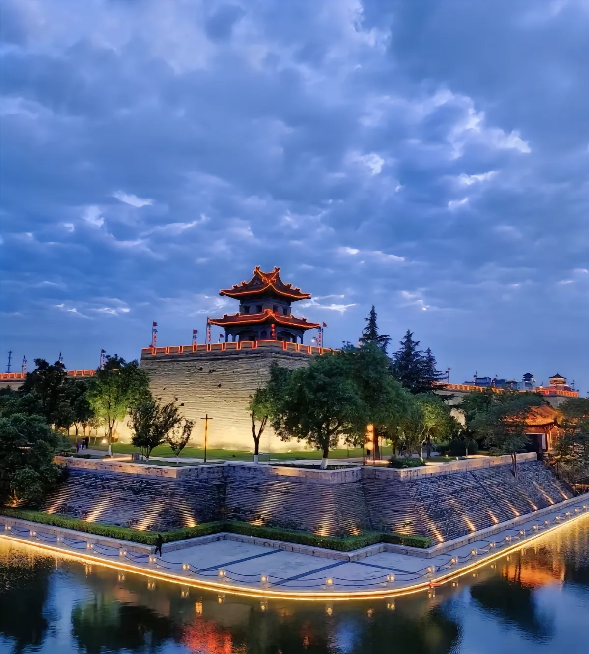 Night view of a traditional Chinese-style pavilion on a hill with trees, surrounded by water, illuminated by streetlights, under a cloudy sky. xi'an city wall