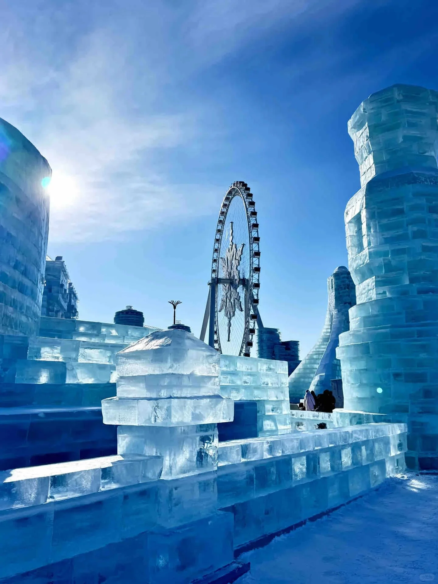 Ice sculpture park with a large Ferris wheel in the background under a clear blue sky.