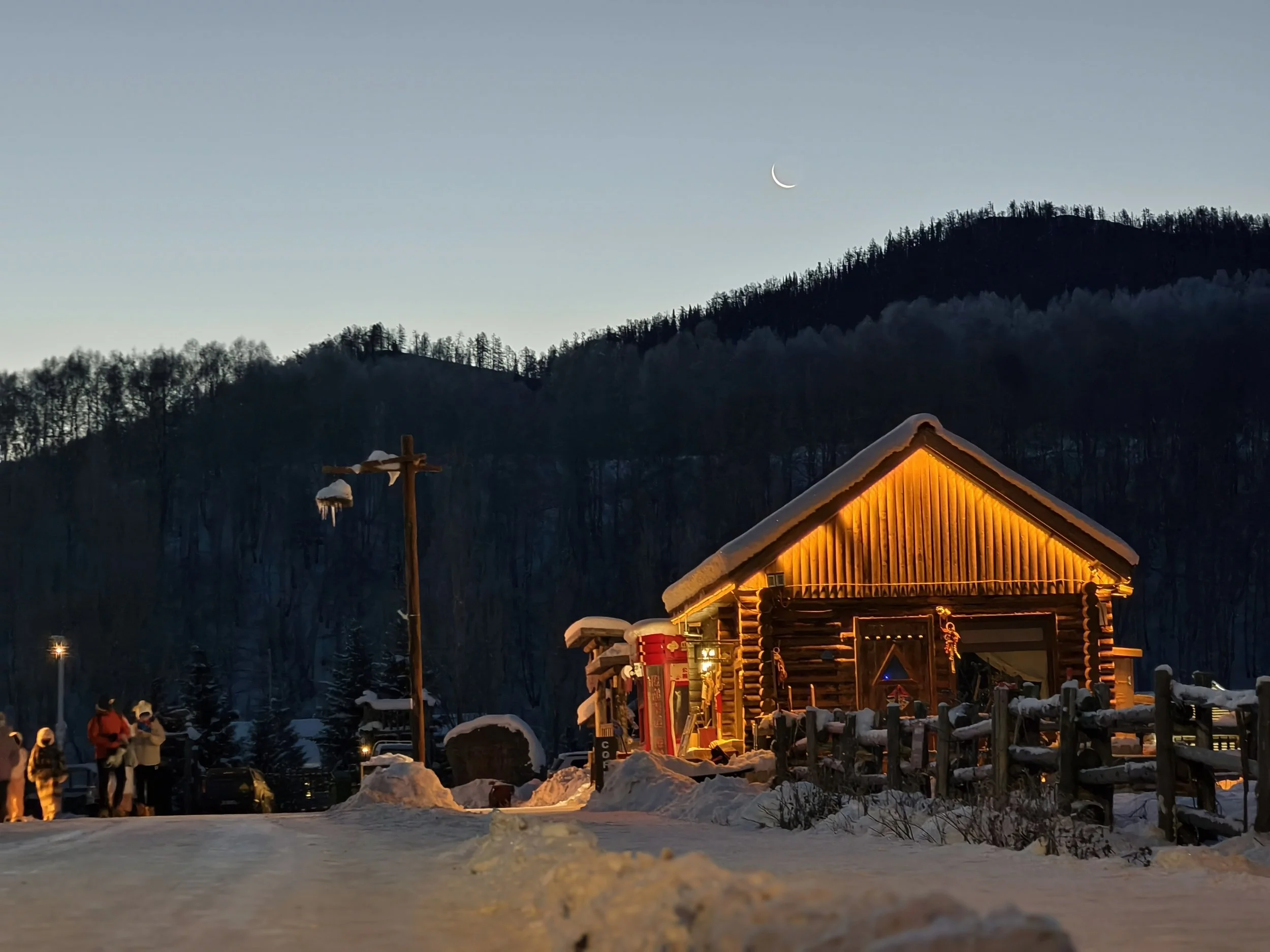 A cozy cabin with warm yellow lights in a snowy landscape at dusk, with a few people standing nearby, a telephone booth, and mountain hills in the background under a sky with a crescent moon, Hemu, Xinjiang.