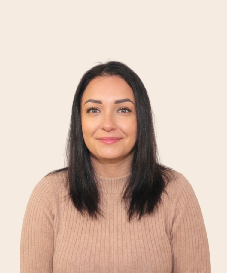 A woman with straight dark hair, light skin, wearing a beige ribbed sweater, smiling, standing against a plain light-colored background.