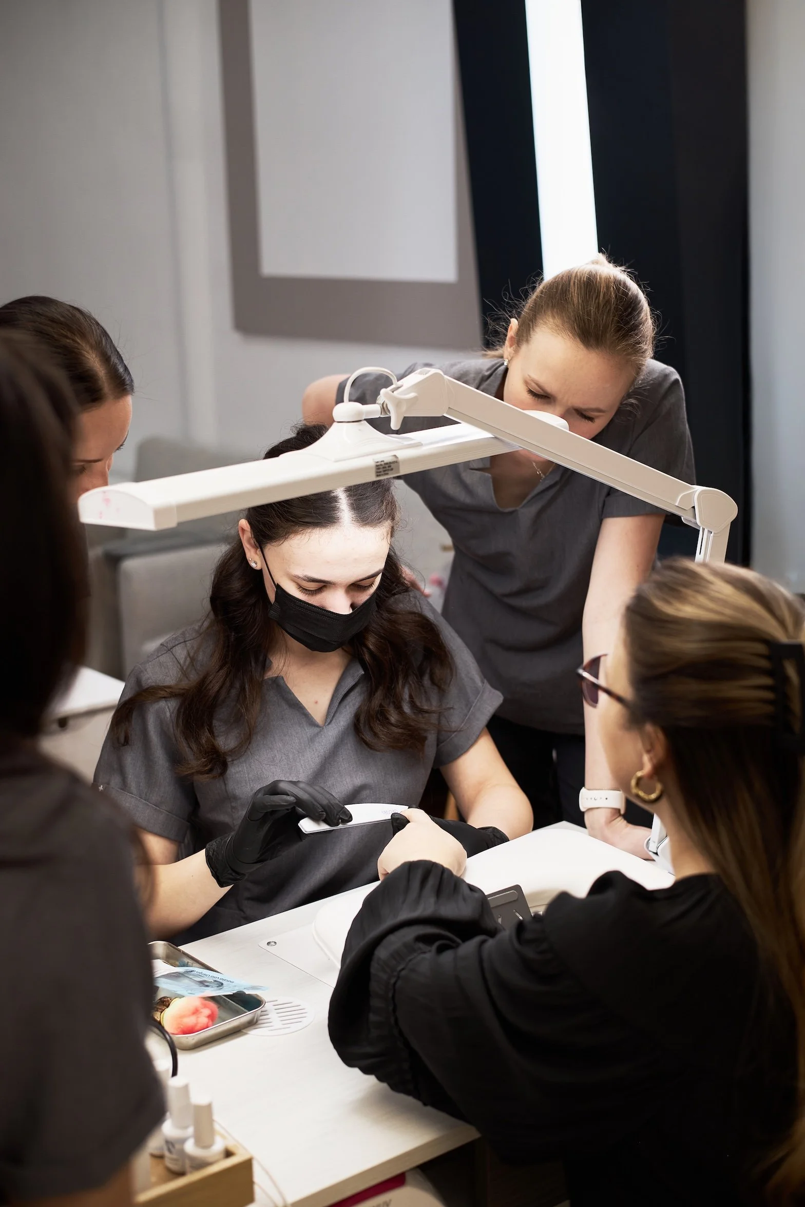 A group of women in dark gray scrubs and face masks working together on a cosmetic procedure, with one woman using a tool and others observing, in a clinical setting.