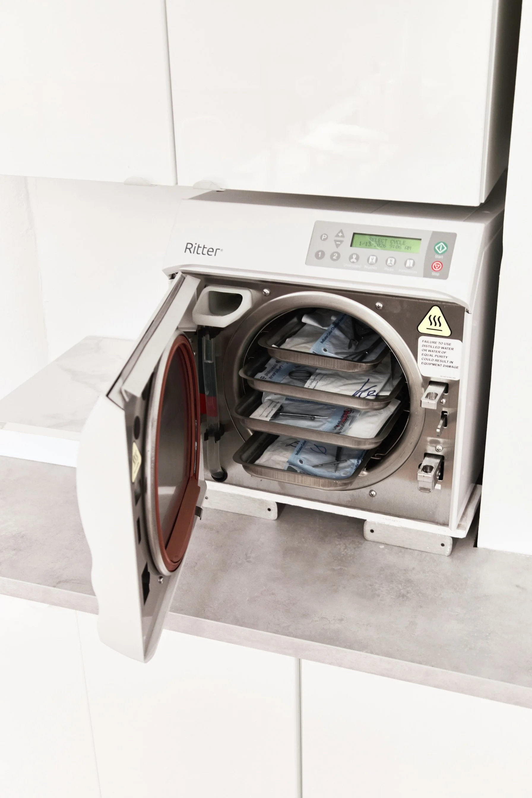 A medical sterilizer with its door open, showing trays of sterilized surgical instruments inside.