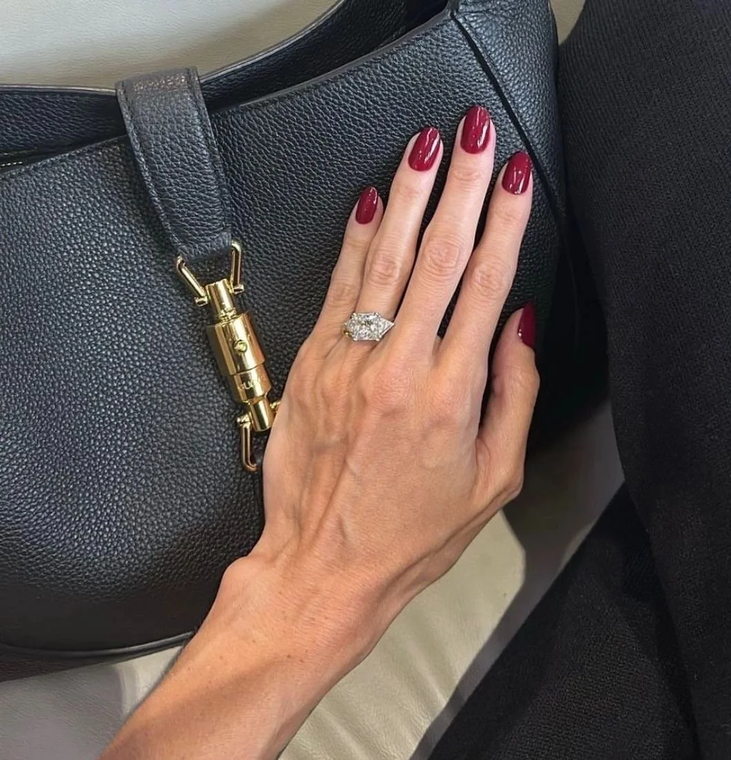 A woman's hand with red nail polish and a large diamond ring resting on a black leather handbag with gold hardware.