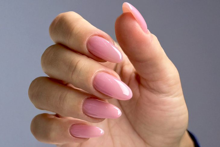 Close-up of a hand with manicured nails painted in a glossy light pink color, set against a gray background.