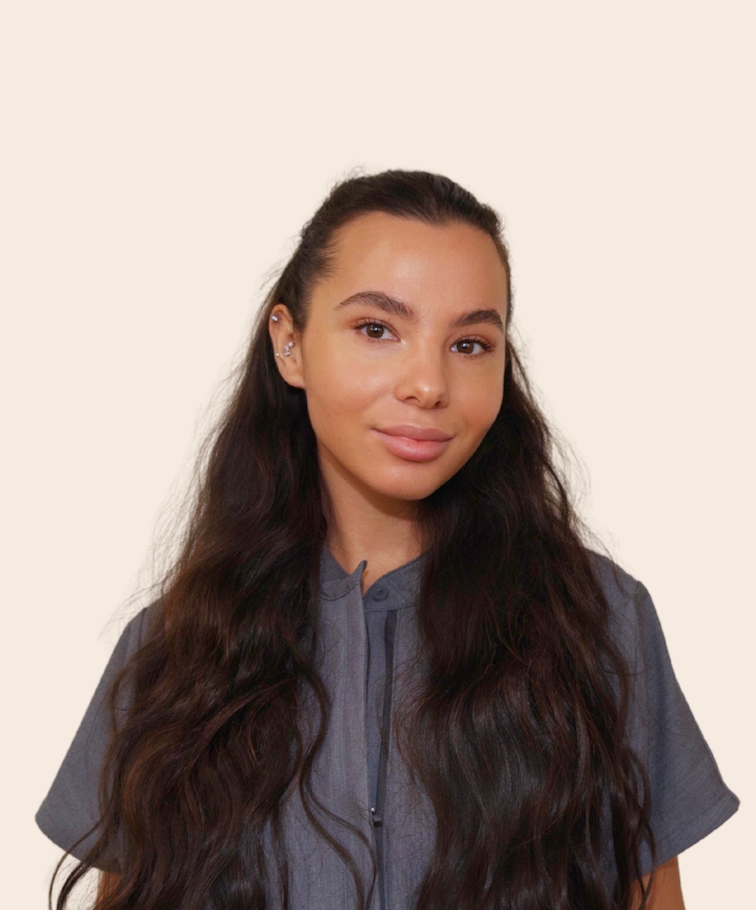 A young woman with long, wavy dark hair and multiple earrings in her left ear, looking at the camera with a neutral expression, wearing a gray shirt against a plain neutral background.