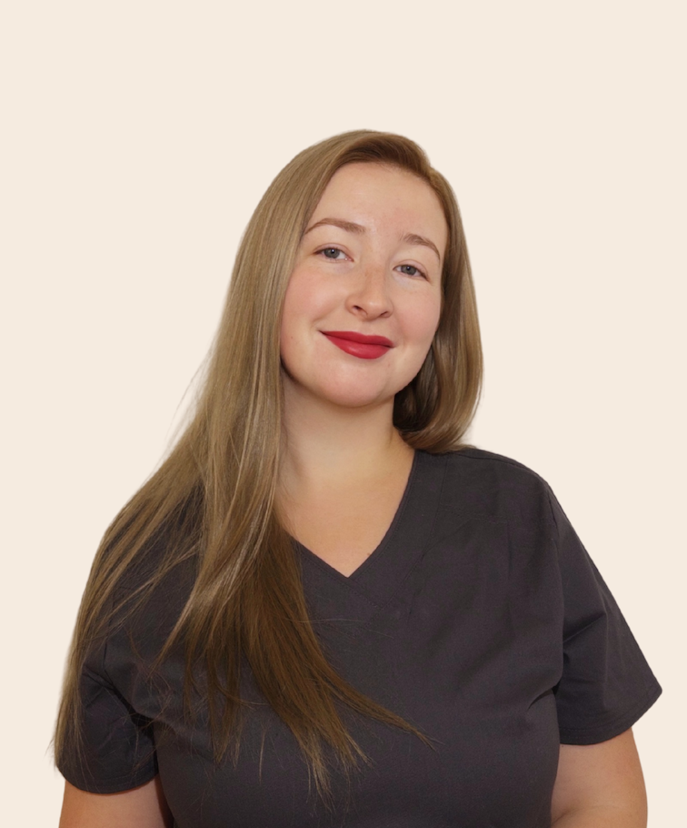 A woman with long, straight, light brown hair and red lipstick, wearing a dark gray medical scrub top, smiling at the camera, against a plain light-colored background.