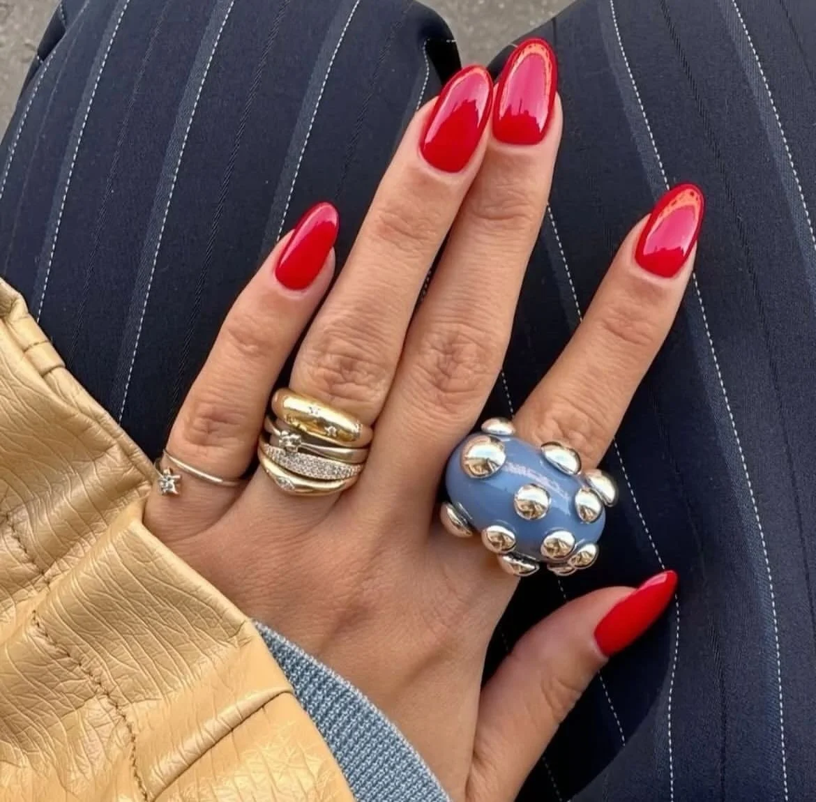 A woman with red painted nails, wearing multiple rings, including a large blue and silver statement ring, resting her hand on her lap with dark pinstriped pants and a tan jacket.