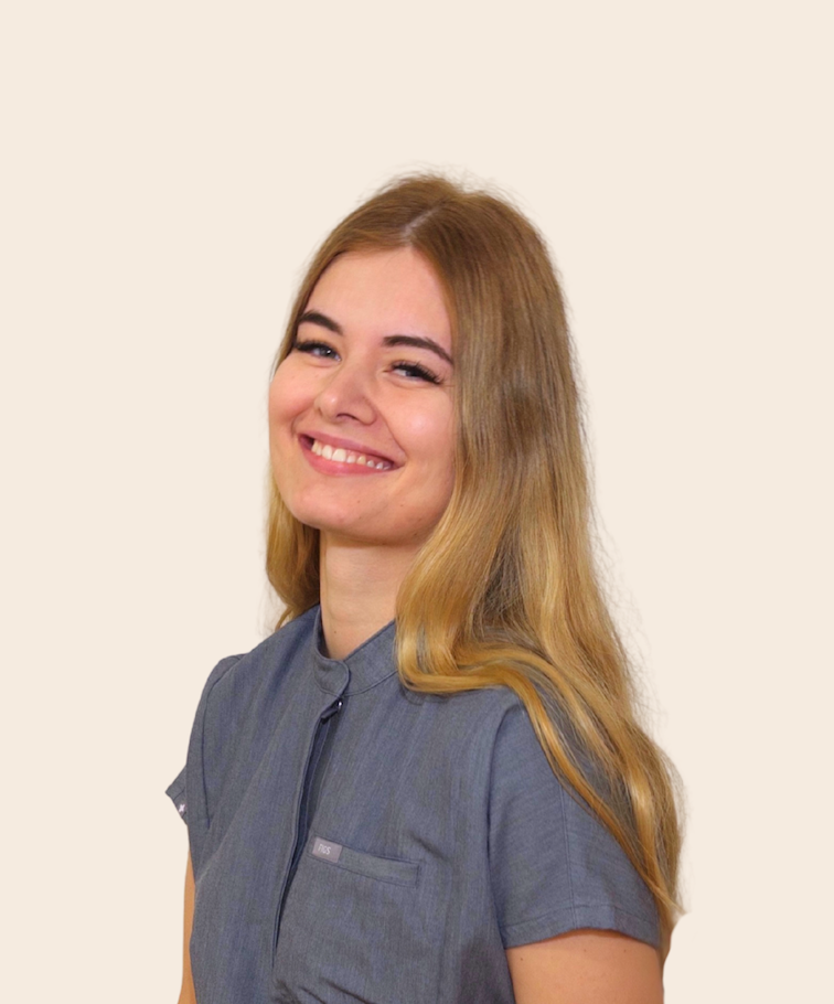 Young woman with long blonde hair smiling, wearing a gray dentist uniform, against a light background.