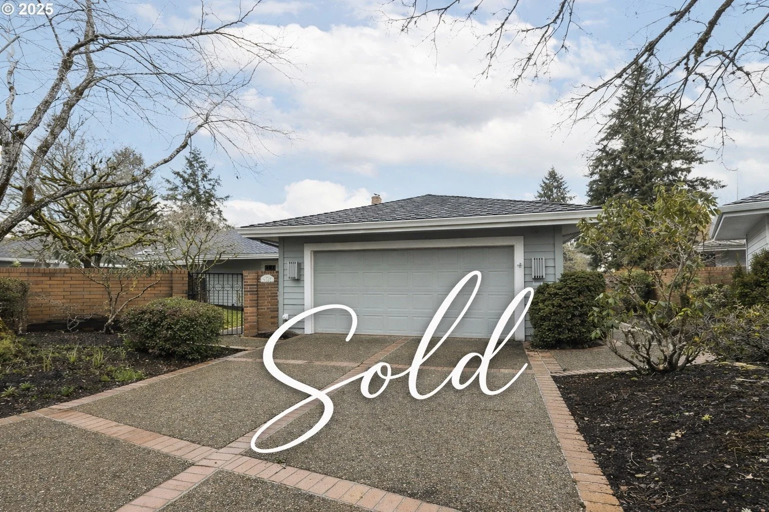 Front view of a house with a driveway, trees, bushes, and a brick wall on the left side. The house has a gray garage door, light blue siding, and a dark gray shingled roof. The word "Sold" is written in large white cursive letters across the image.