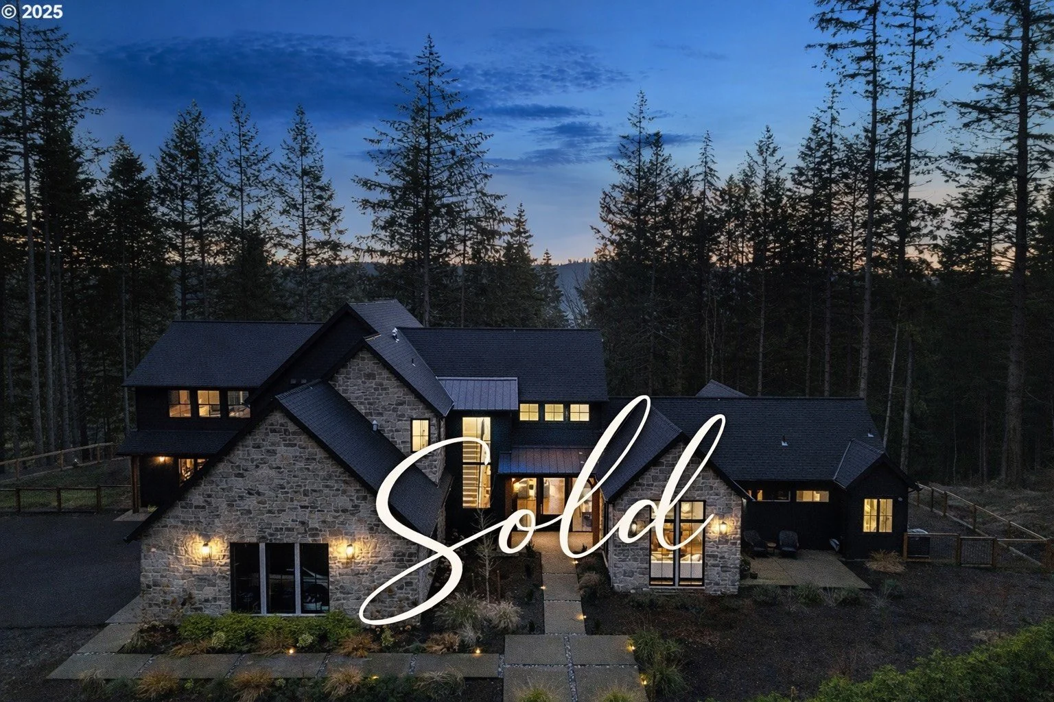A modern house made of stone and dark siding with multiple pitched roofs, large windows, and outdoor lighting, surrounded by tall trees at dusk with a partly cloudy sky in the background. The word "Sold" is overlaid in cursive.