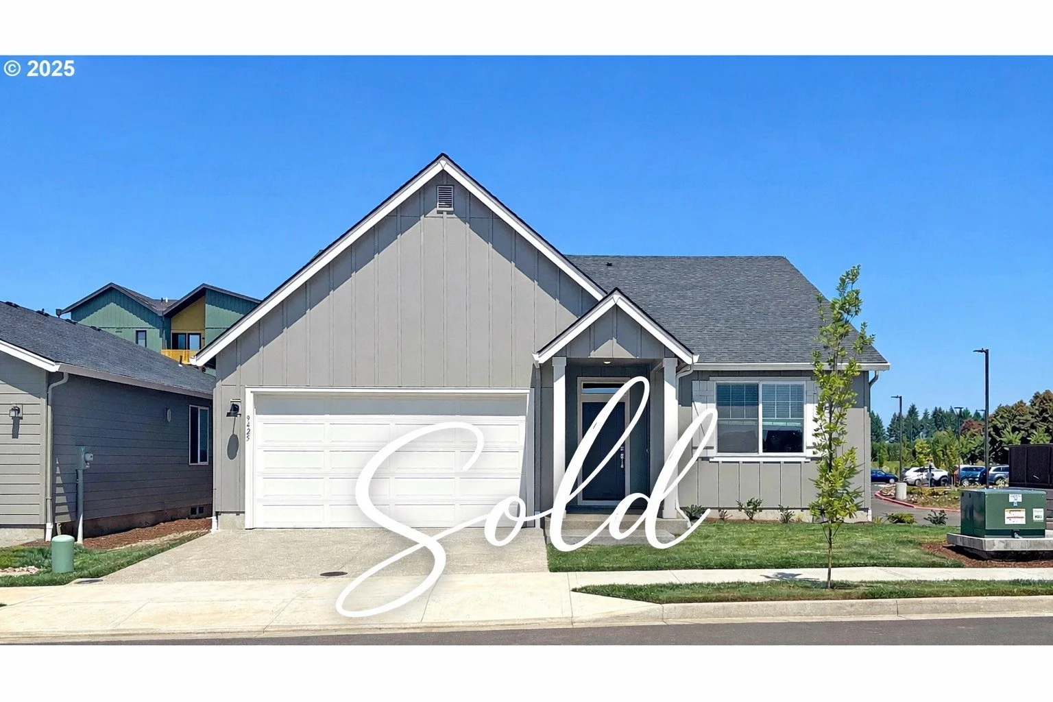 A modern suburban house with a gray exterior, white garage door, small front lawn, and young tree, under a clear blue sky with the word "Sold" overlayed.