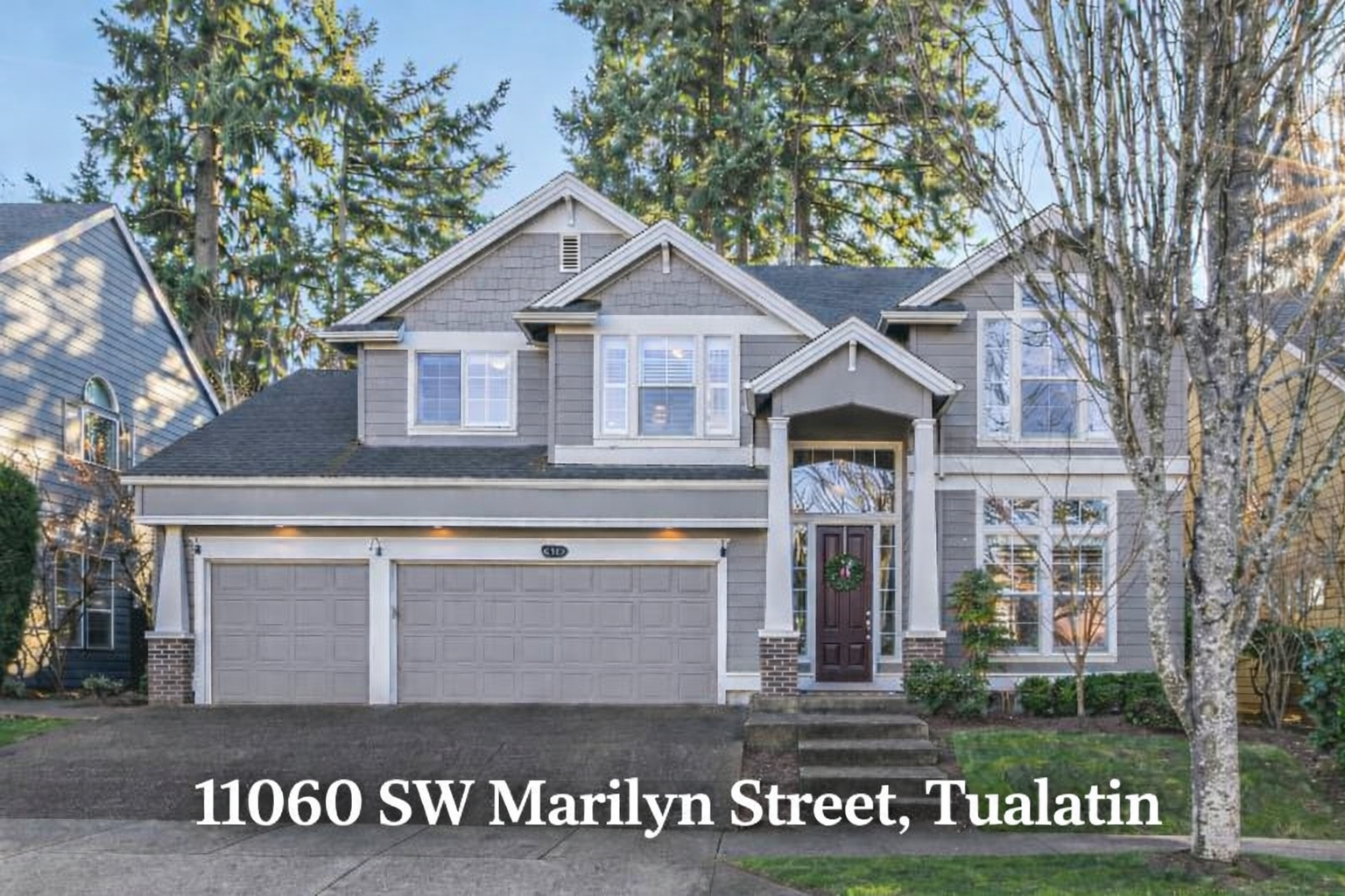 Front view of a two-story house with a gray exterior, white trim, and a dark wood front door decorated with a green wreath, situated at 11060 SW Marilyn Street, Tualatin. The house has a two-car garage, multiple windows, and is surrounded by trees and greenery.