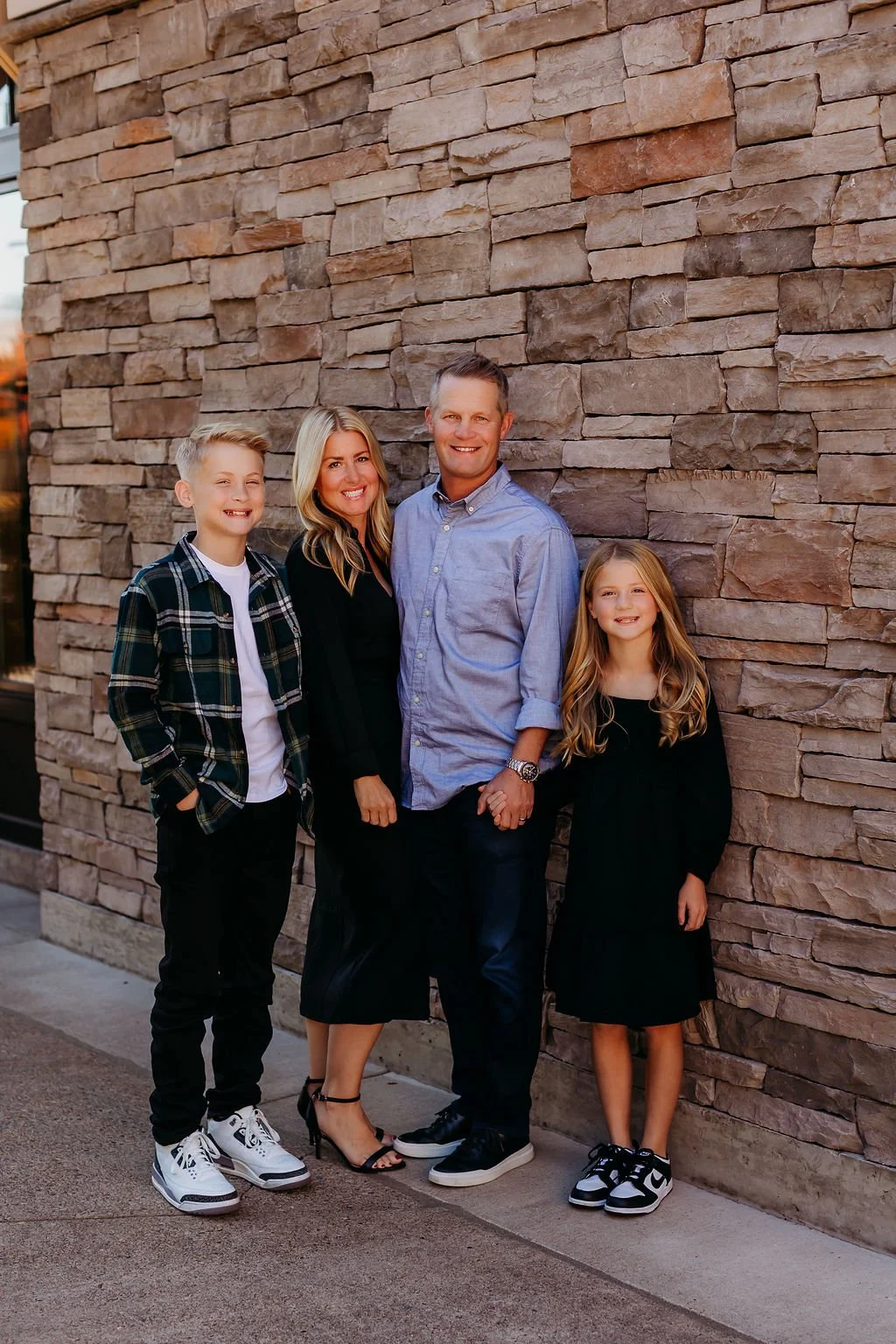 A family of four standing in front of a beige brick wall, smiling at the camera. The family includes a boy, a woman, a man, and a girl, all dressed in casual and semi-formal clothing.