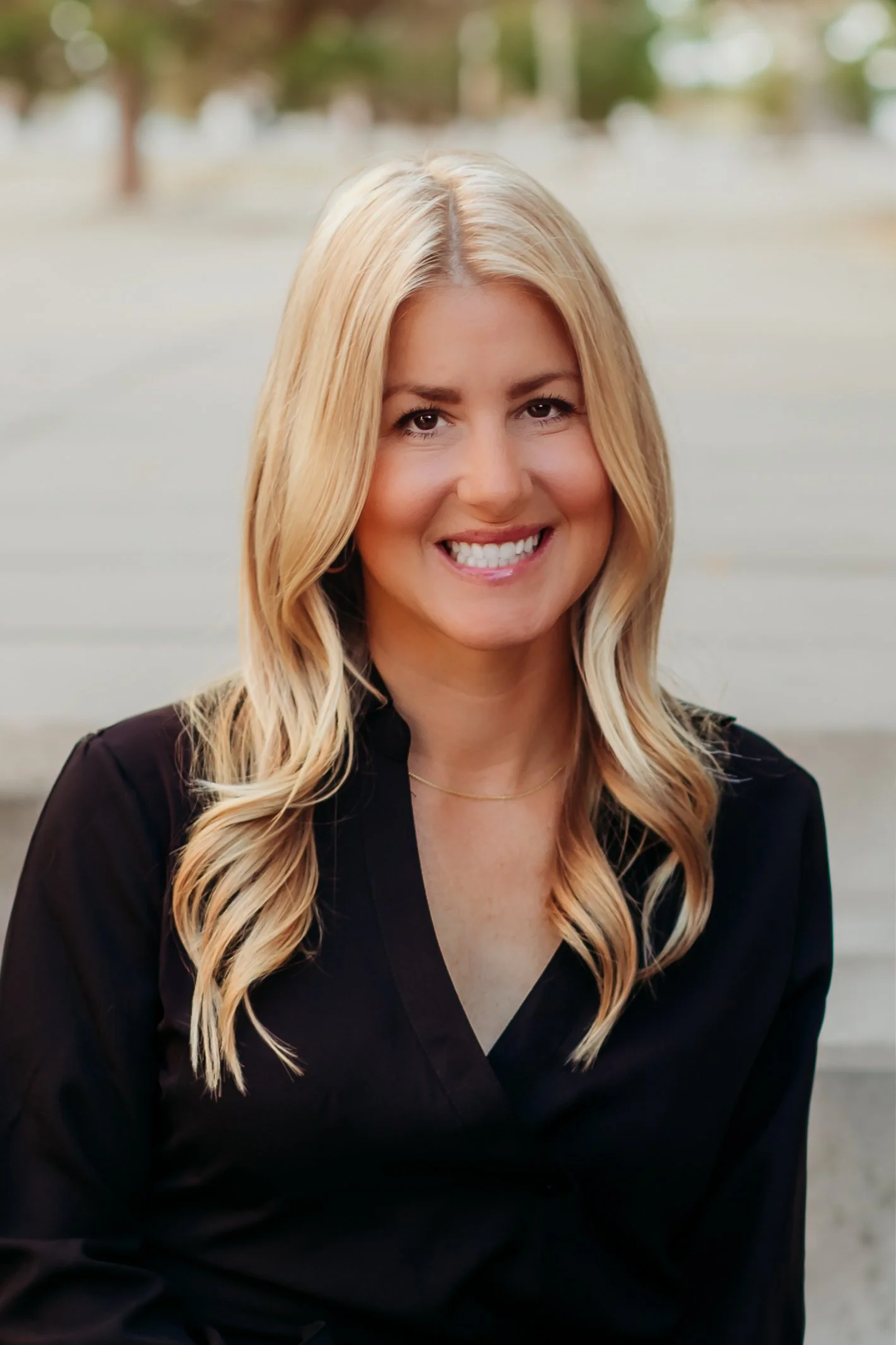 A blonde woman with long, wavy hair smiling in an outdoor setting.