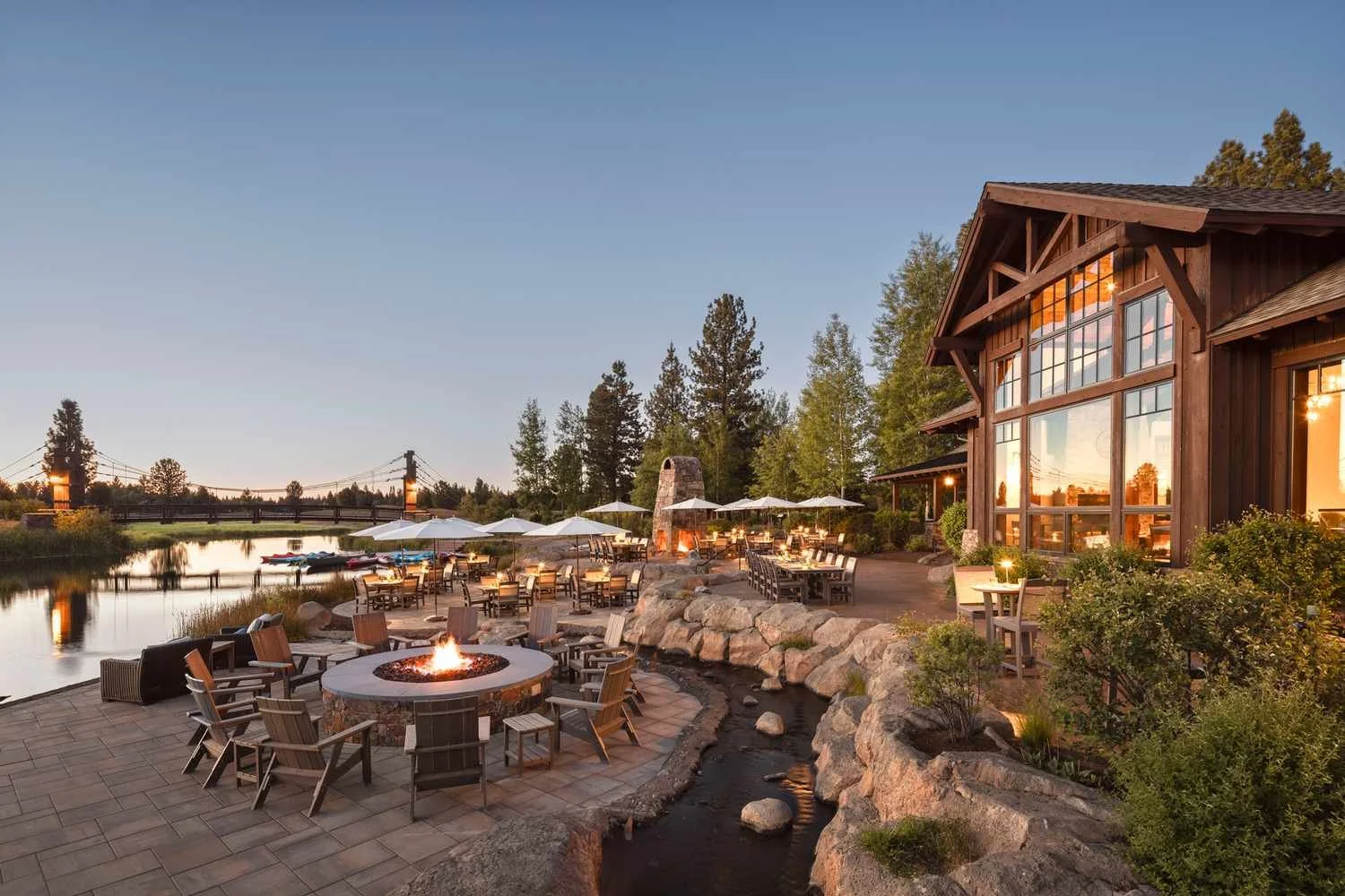 Outdoor patio area at sunset with a fire pit, tables with umbrellas, and a large wooden building, overlooking a calm body of water with trees in the background.